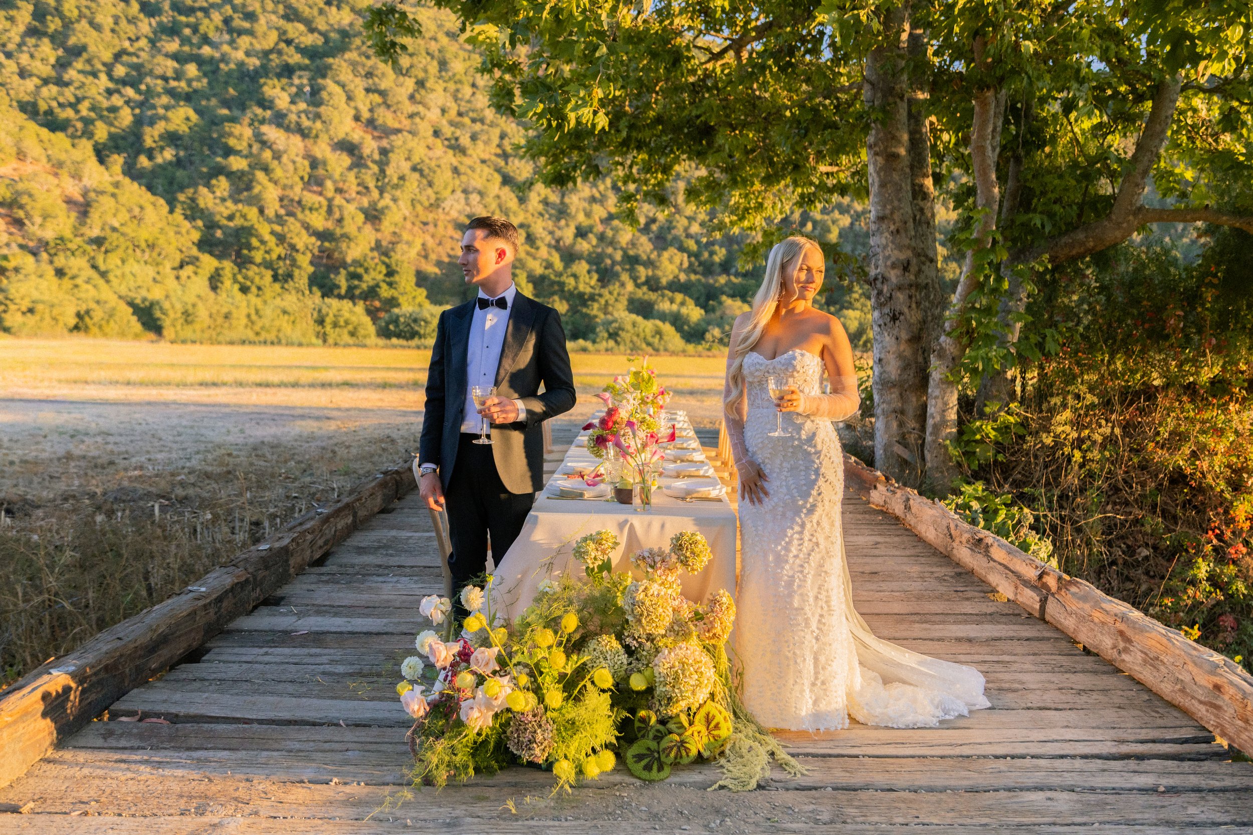 A bride and groom standing outdoors on a wooden bridge, holding glasses of wine, with a long table decorated with flowers and surrounded by trees and mountains in the background during sunset.