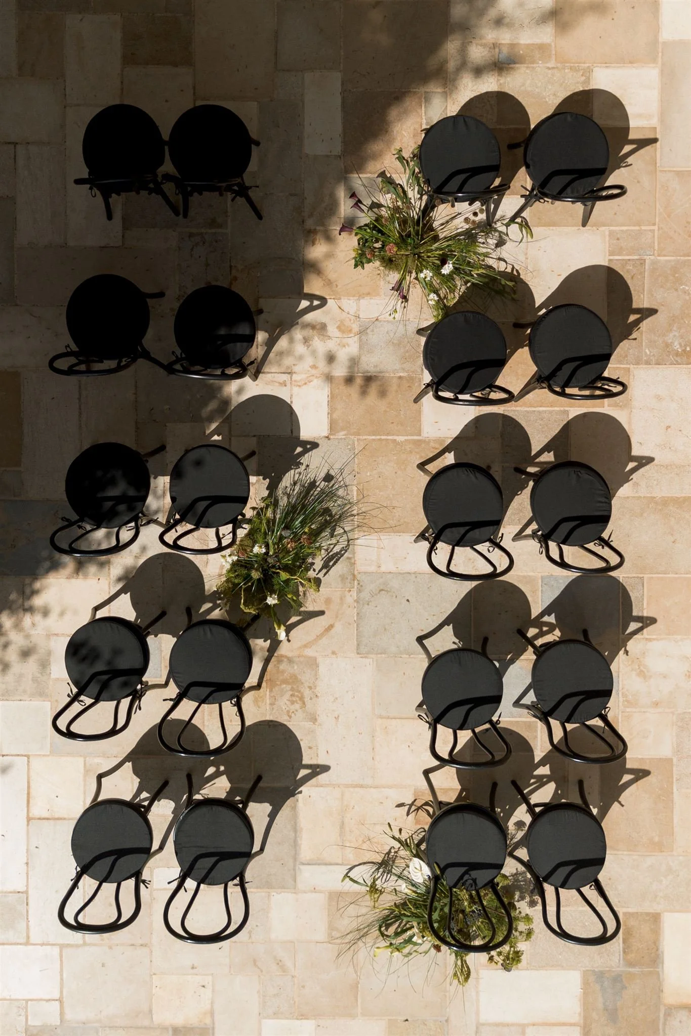 Top-down view of nine black chairs arranged in three rows around three circular tables with flower arrangements in the center, on a tiled floor with sunlight casting shadows.