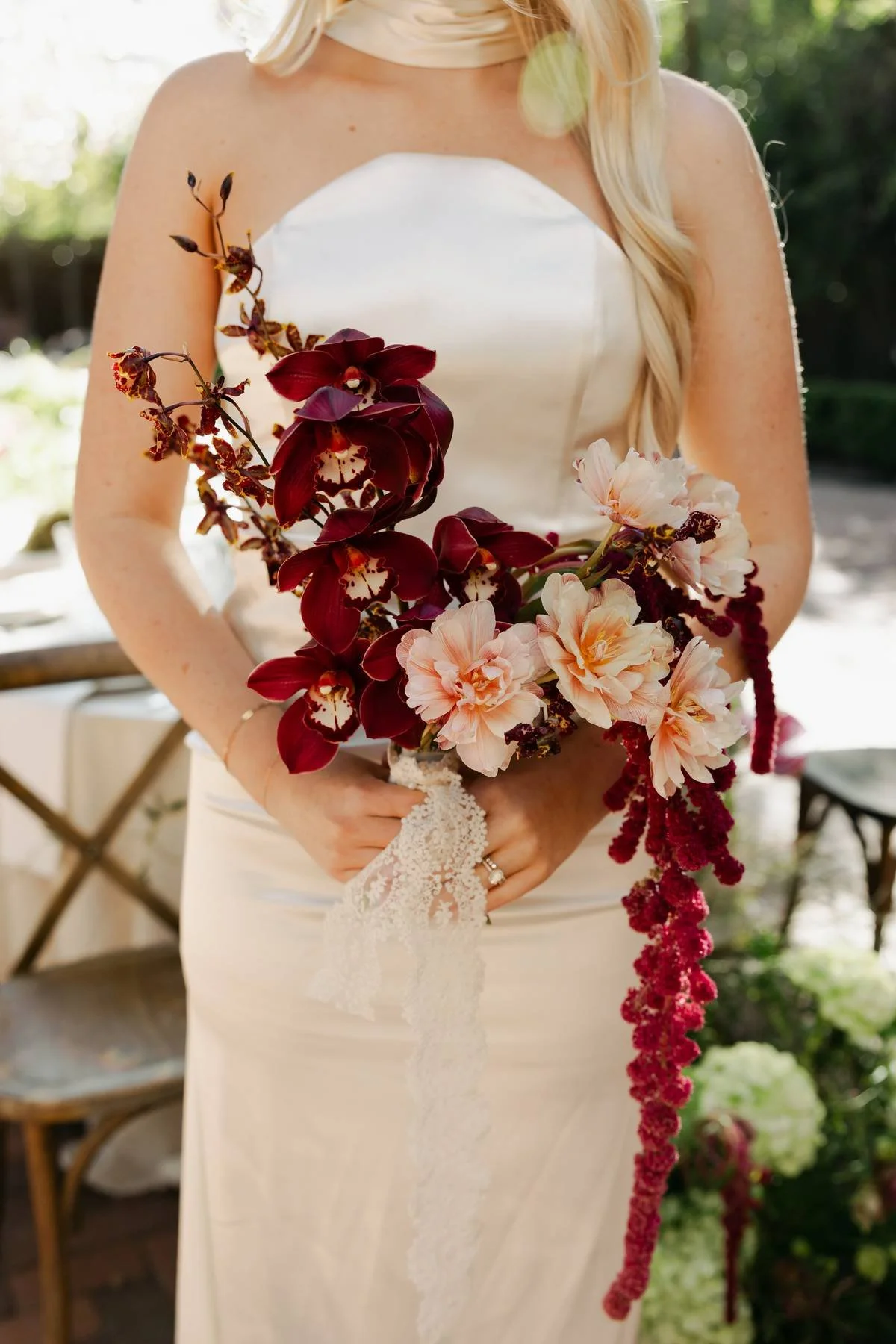 A bride in a white strapless dress holding a bouquet of pink and burgundy flowers, with lace gloves, outdoors with greenery in the background.