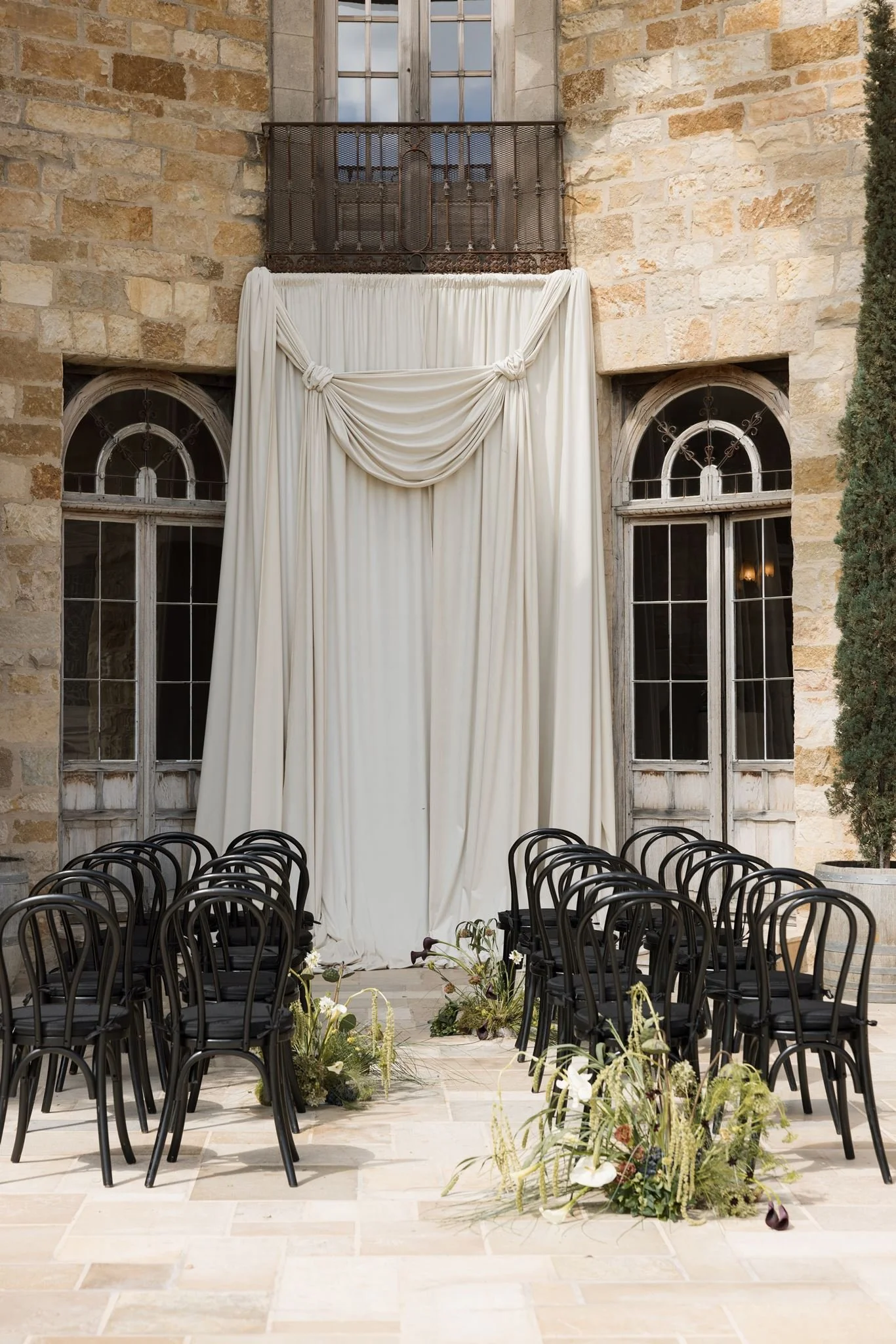 An outdoor wedding ceremony setup with black chairs arranged in rows facing a white draped fabric backdrop. The backdrop is decorated with greenery and flowers. Behind the backdrop, there are two arched windows in a stone building, with a stone wall 