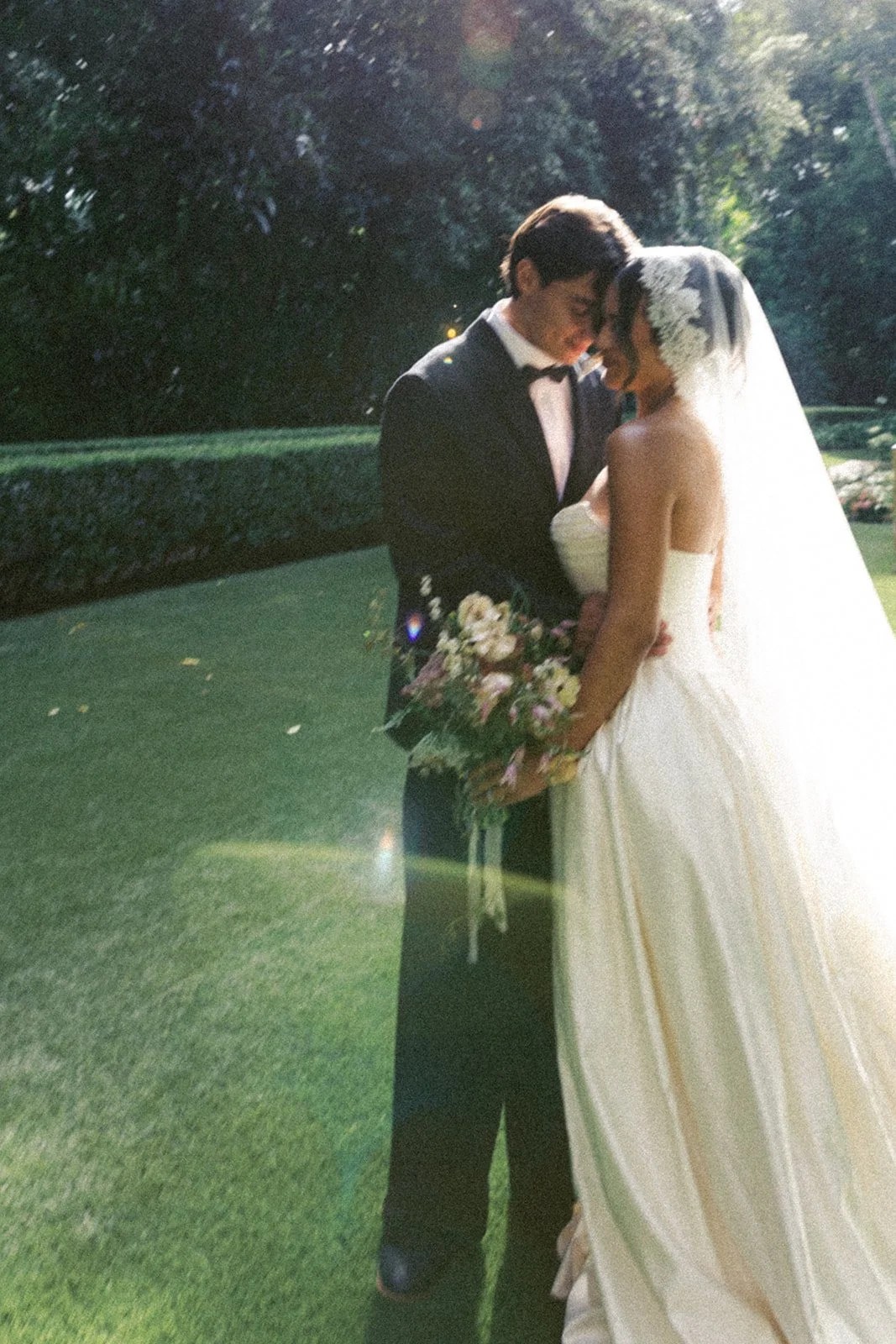 A bride and groom stand close together outdoors on grass, touching foreheads and smiling with eyes closed, during their wedding. The groom wears a tuxedo, and the bride wears a strapless wedding gown, a veil, and holds a bouquet of flowers. Sunlight filters through trees in the background.