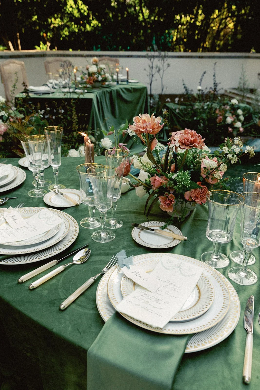 Elegant outdoor dining table with green tablecloth, floral centerpiece, white and gold china, silverware, glassware, and handwritten place cards, set against a backdrop of greenery and additional tables with floral decor.