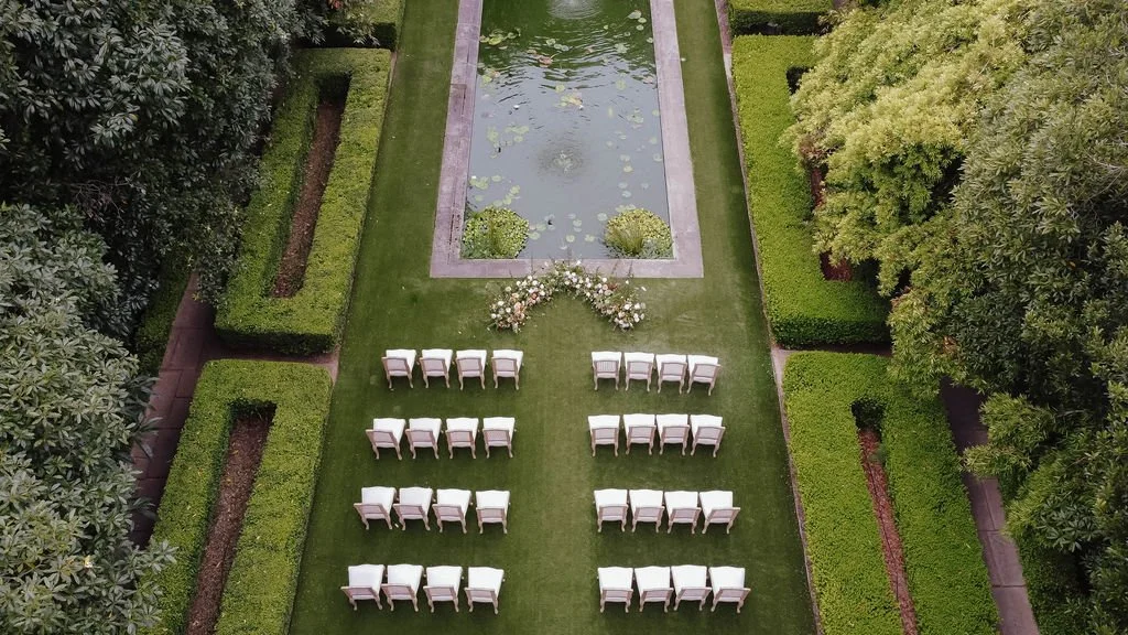An outdoor wedding setup with white chairs arranged in rows on green grass facing a decorative arch. There is a small pond with lily pads in the background, surrounded by well-manicured hedges and trees.