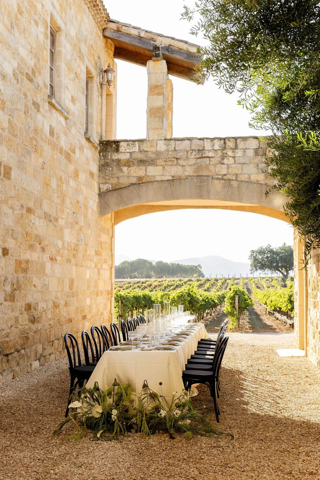 A long dining table set with white tablecloths, glassware, and plates under a stone archway overlooking a vineyard at sunset.