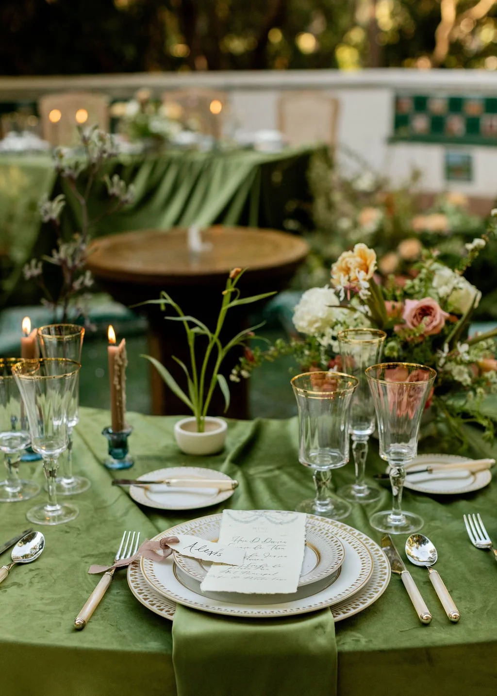 Elegant outdoor dining table with gold-rimmed plates, silverware wrapped with ribbons, floral centerpiece, candles, and glassware, set for a formal event in a garden or patio setting.