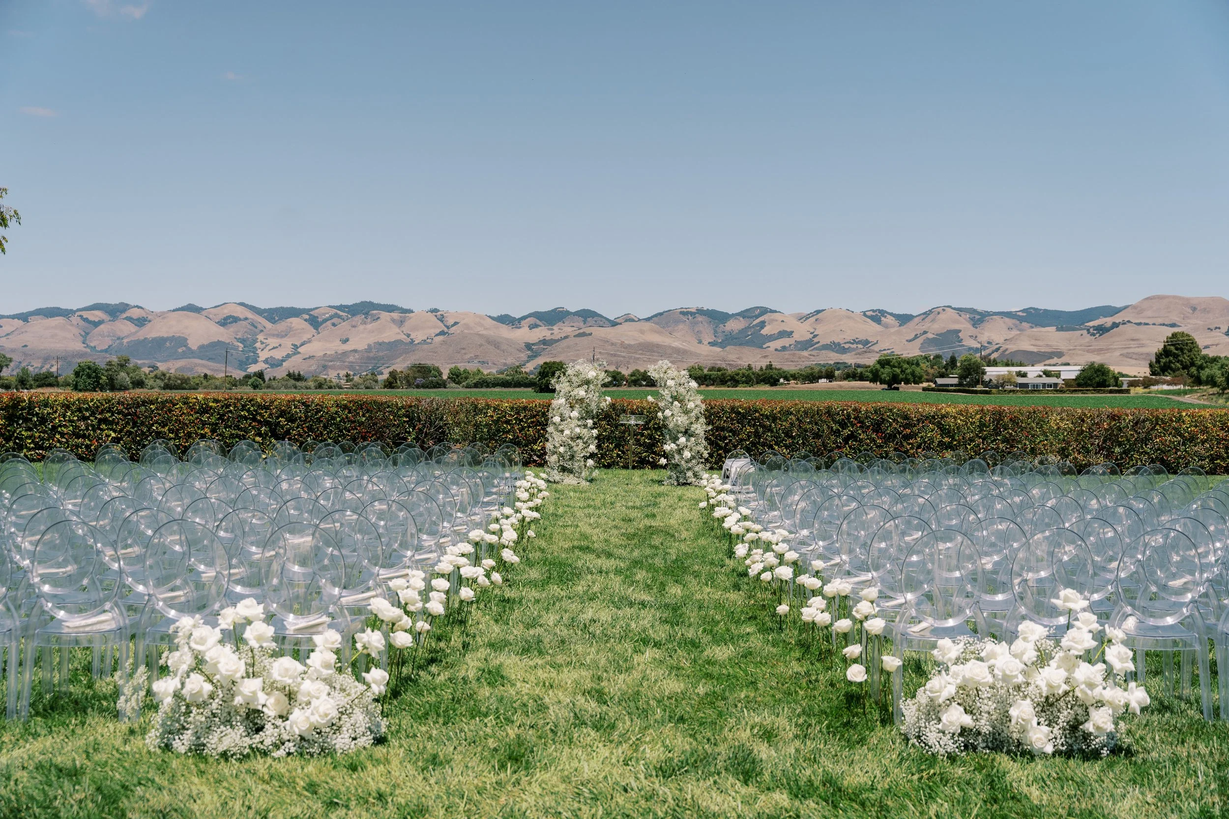 Outdoor wedding setup on a green lawn with transparent chairs arranged in rows, white floral decorations along a central aisle, and mountains in the background under a clear blue sky.
