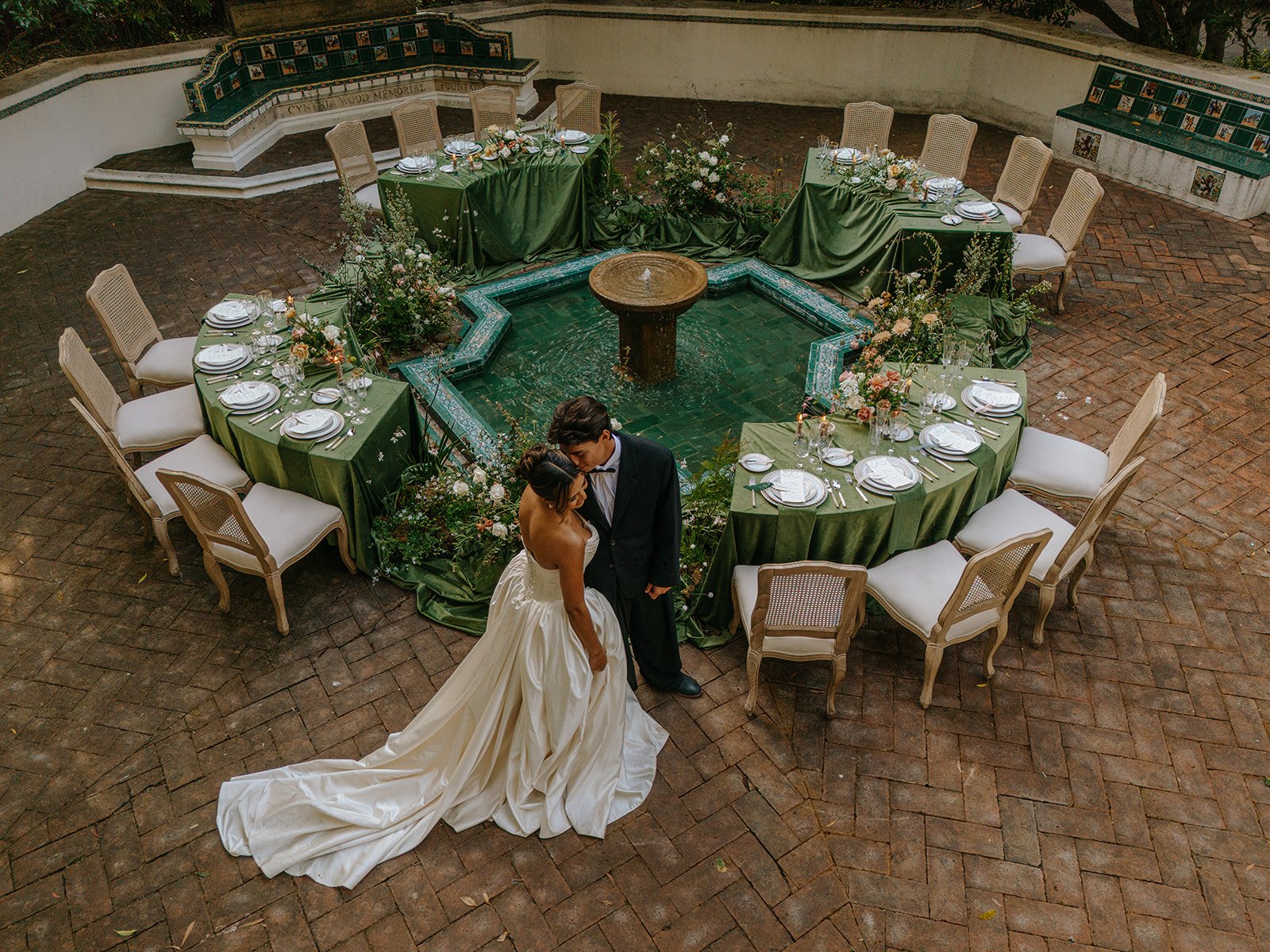A bride and groom standing close together near a fountain, surrounded by a circular arrangement of decorated tables set for a wedding reception, with white chairs and floral centerpieces.