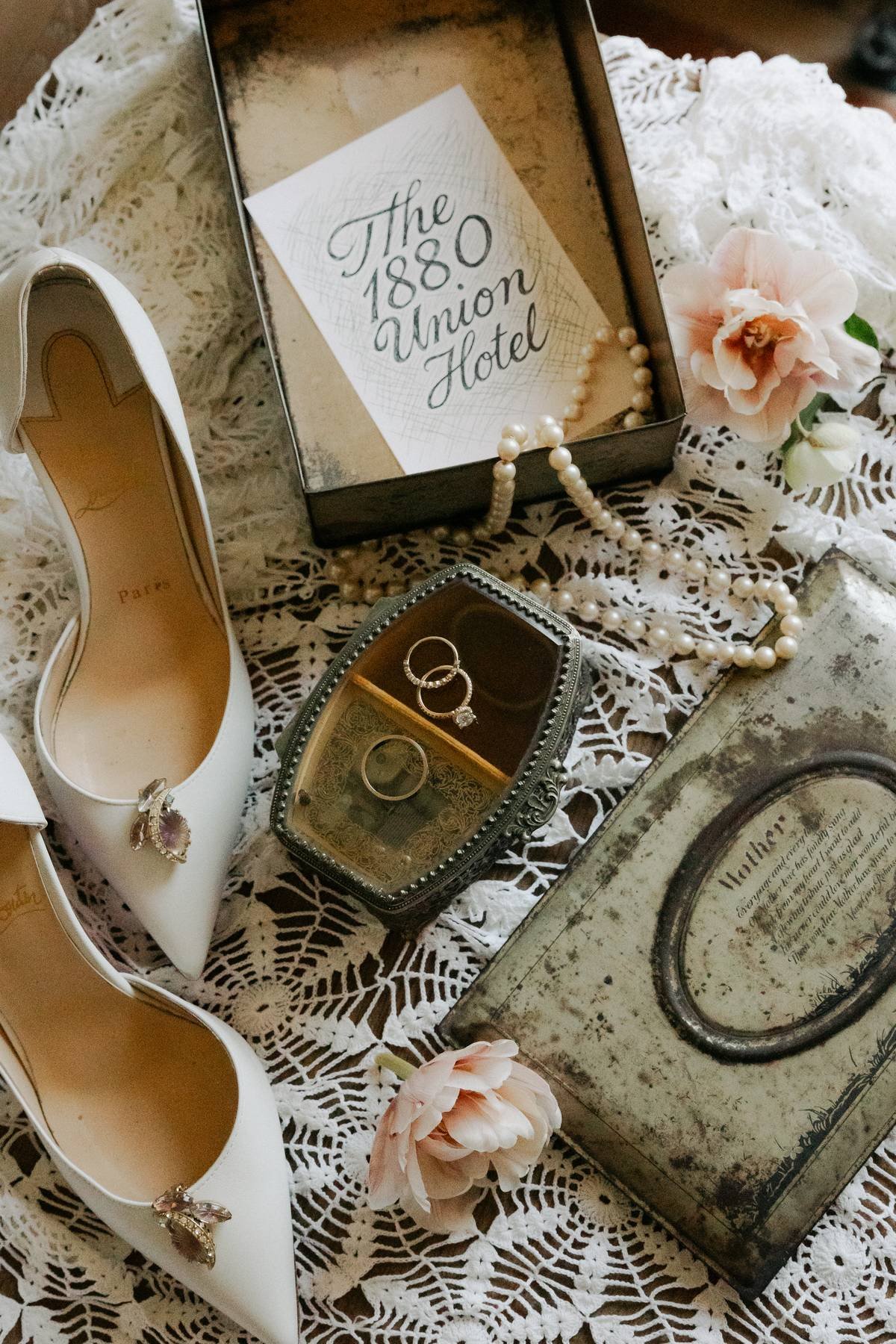 A vintage jewelry box with pearl necklace and rings, white high heel shoes with rings, a lace cloth, pink flowers, and a decorative box with the text 'The 1880 Union Hotel' on it.