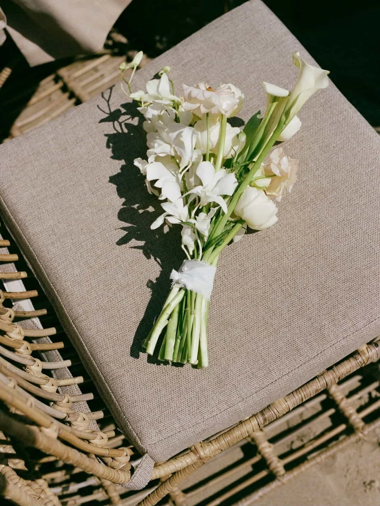 A bouquet of white flowers, including calla lilies, arranged and tied with a white ribbon, resting on a beige cushion on a wicker chair.
