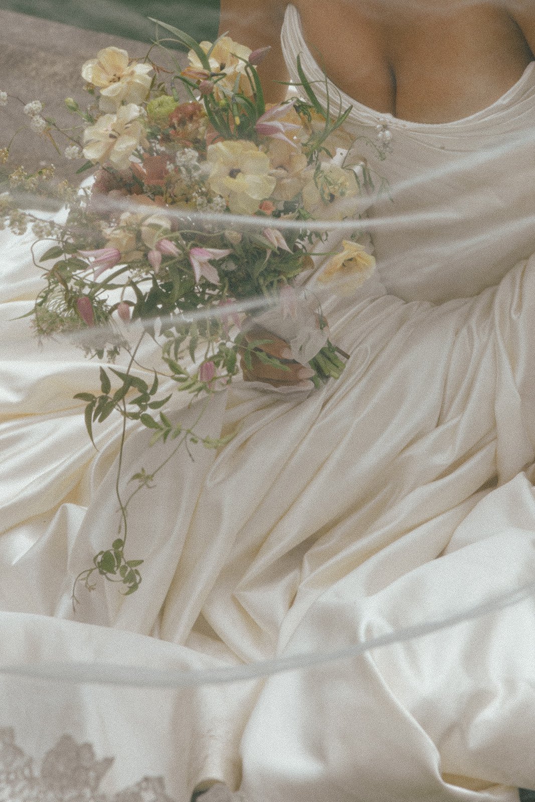 A bride holding a bouquet of flowers, wearing a white satin wedding dress with a low neckline, against a blurred background.