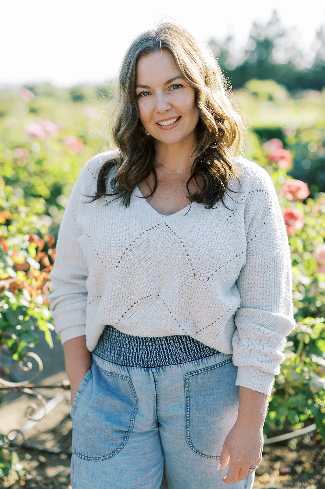 A woman with shoulder-length wavy brown hair, smiling, standing outdoors in a garden with pink flowers, wearing a white sweater and light blue jeans.