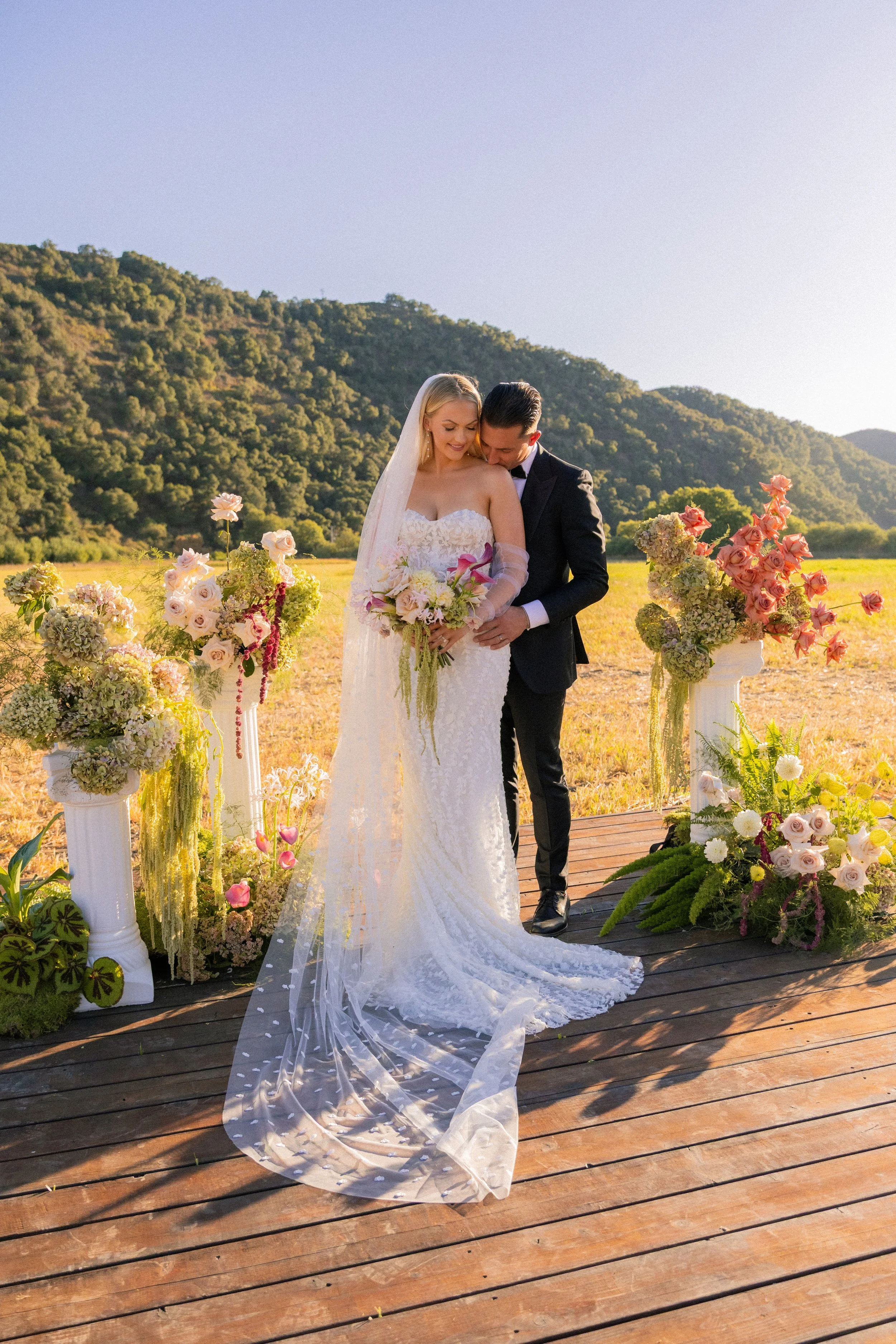 A bride and groom standing on a wooden platform outdoors, surrounded by floral arrangements, with green hills in the background during sunset.