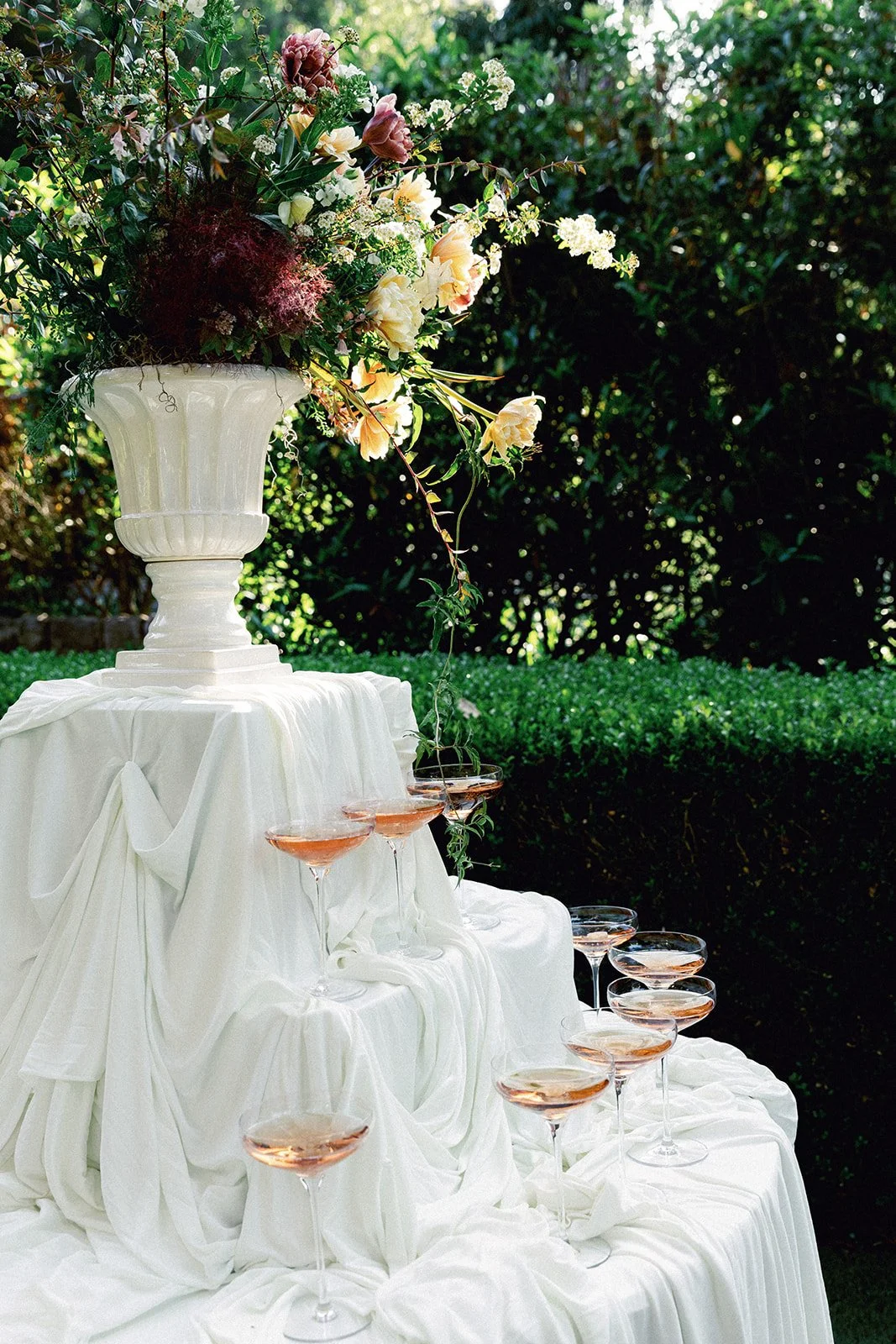 Elegant outdoor table setup with a white draped tablecloth, a large white vase filled with pink, white, and purple flowers, and multiple glasses of pink champagne arranged on a tiered stand, set against green foliage.