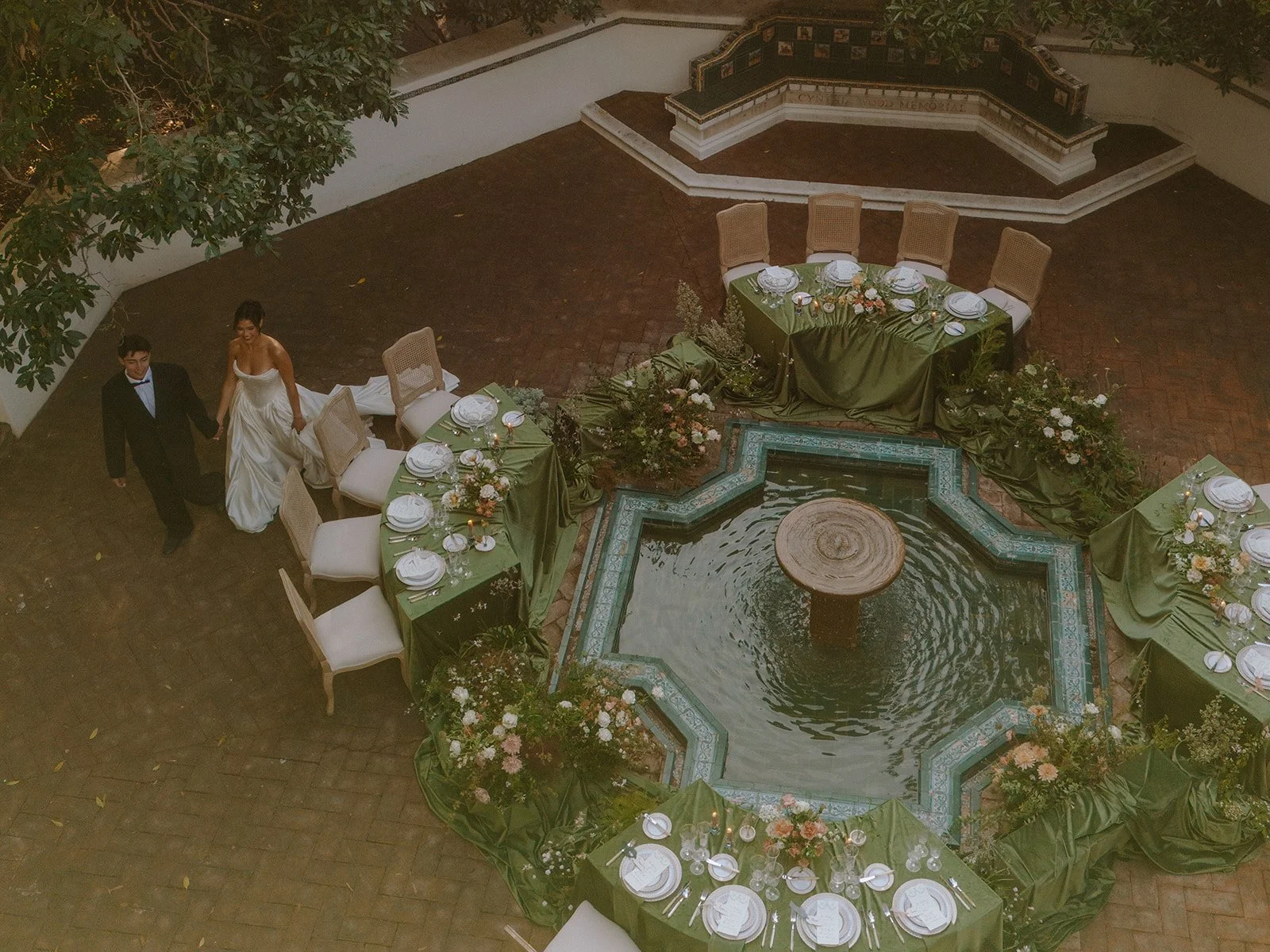 A wedding reception setup with tables decorated with green tablecloths, floral arrangements, and plates, arranged around a small fountain with a water feature in the courtyard. A bride and groom are walking hand in hand.