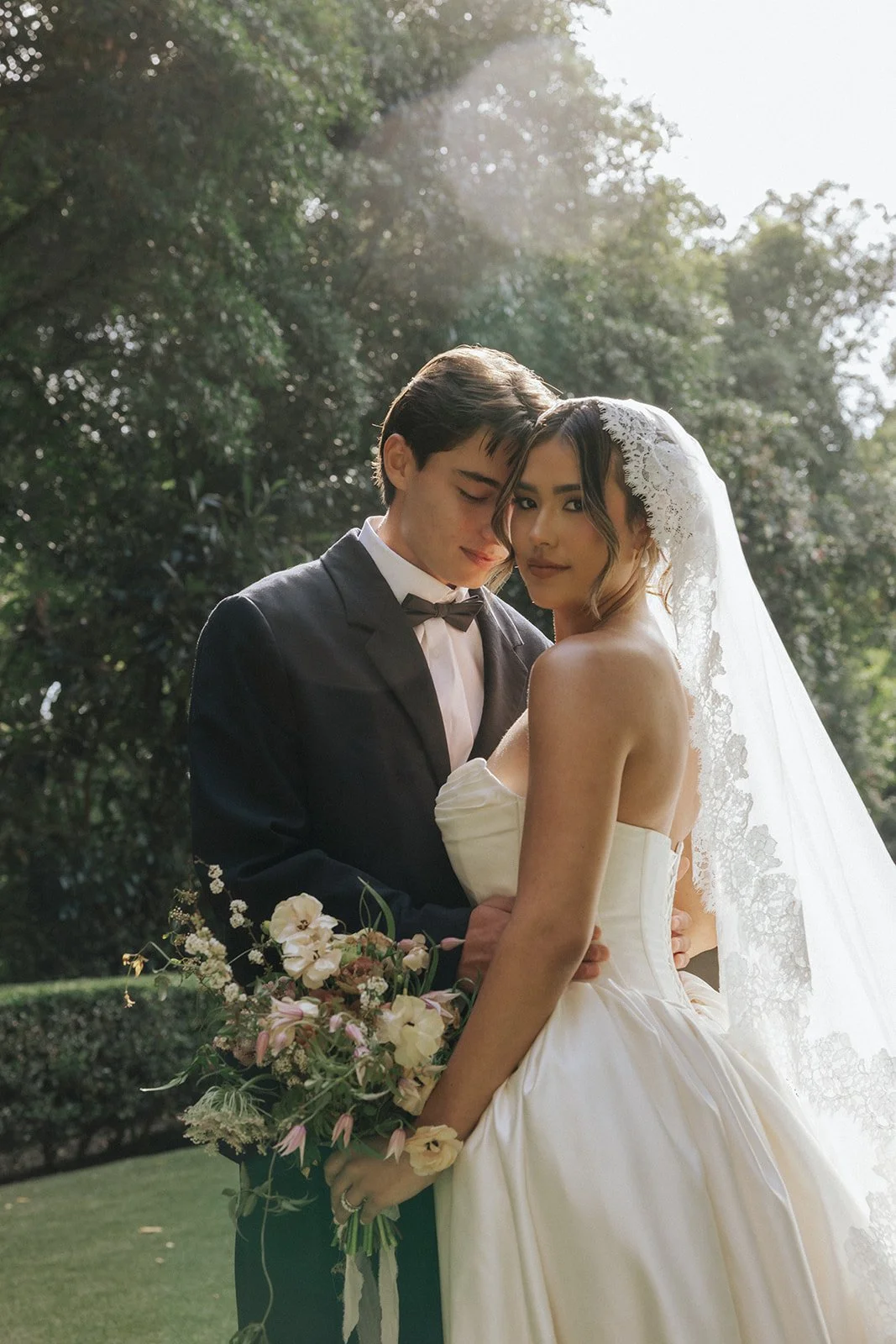 A newlywed couple stands outdoors in a garden, with the groom in a tuxedo and the bride in a white strapless wedding dress with lace veil, holding a bouquet of flowers, standing close with their foreheads touching and eyes closed.