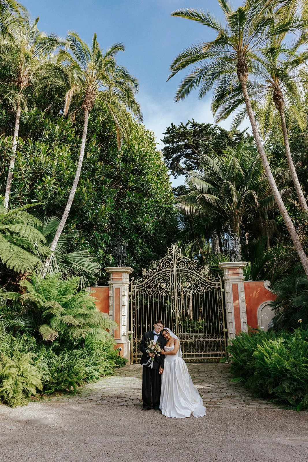 A bride and groom standing in front of a decorative iron gate surrounded by lush tropical plants and tall palm trees on a bright, sunny day.