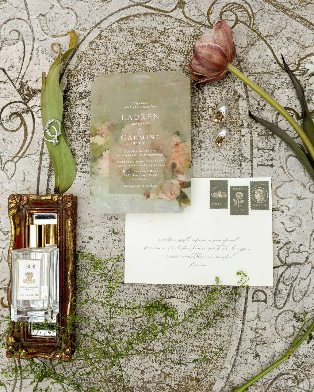 Wedding invitation, vintage perfume bottle, earrings, tulip flower, leaf, and postage card arranged on a textured surface with floral design.