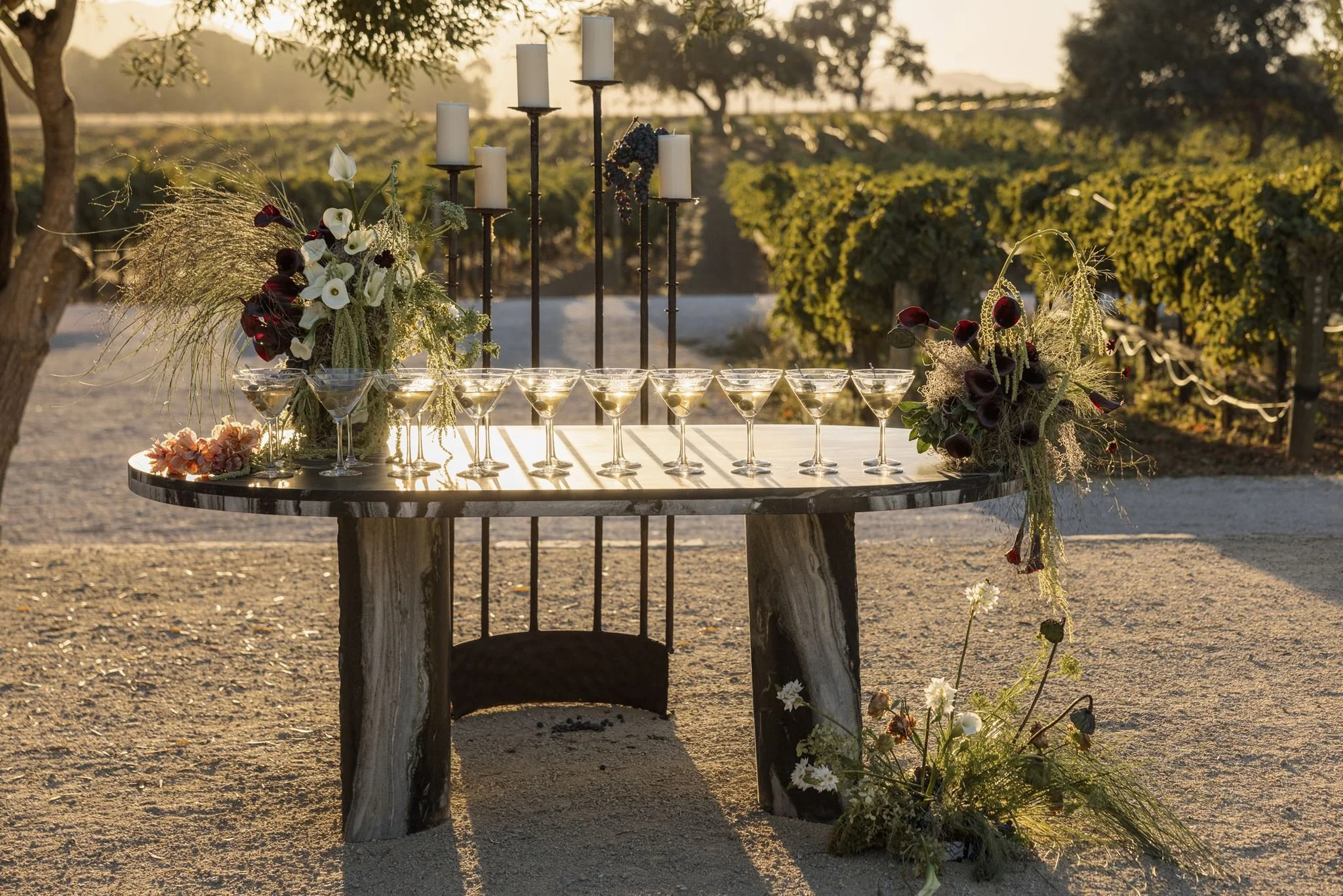 A rustic outdoor table decorated with white and dark red flowers, candles, and glasses of a clear beverage, set in a vineyard at sunset.