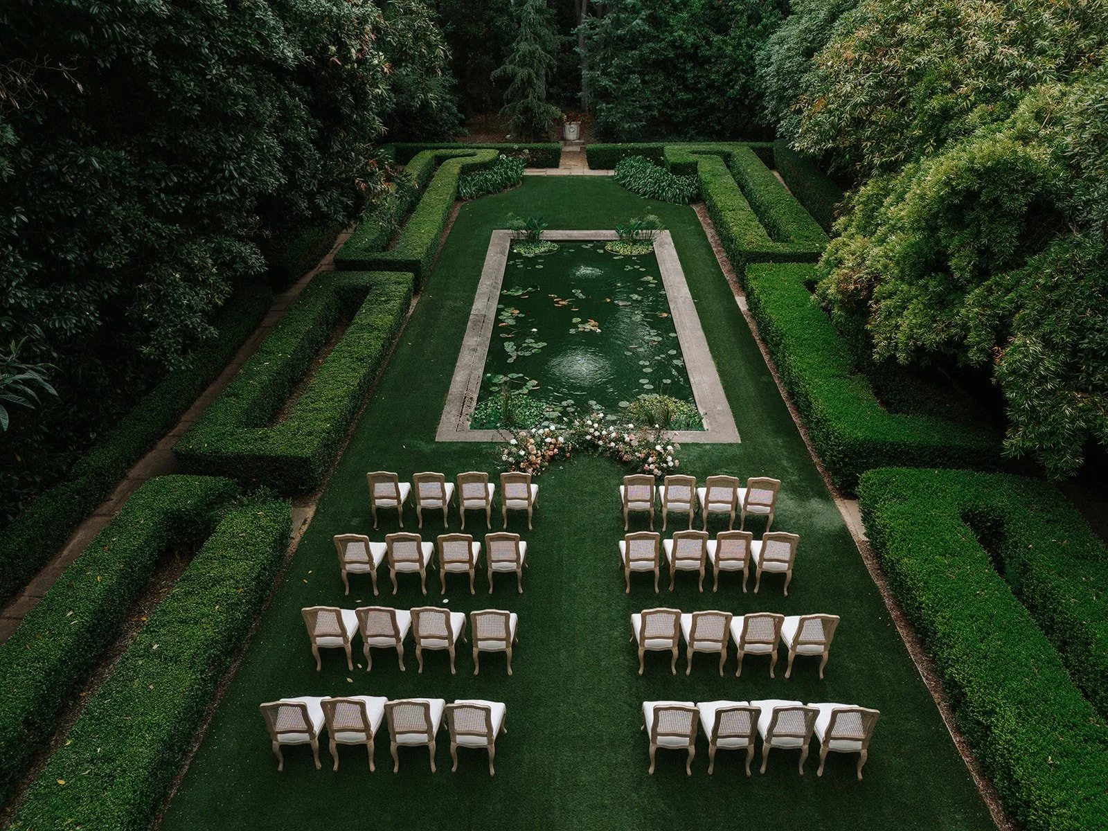 A wedding ceremony setup in a lush garden with white chairs arranged in rows facing a small pond decorated with water lilies, surrounded by well-manicured green hedges and trees.
