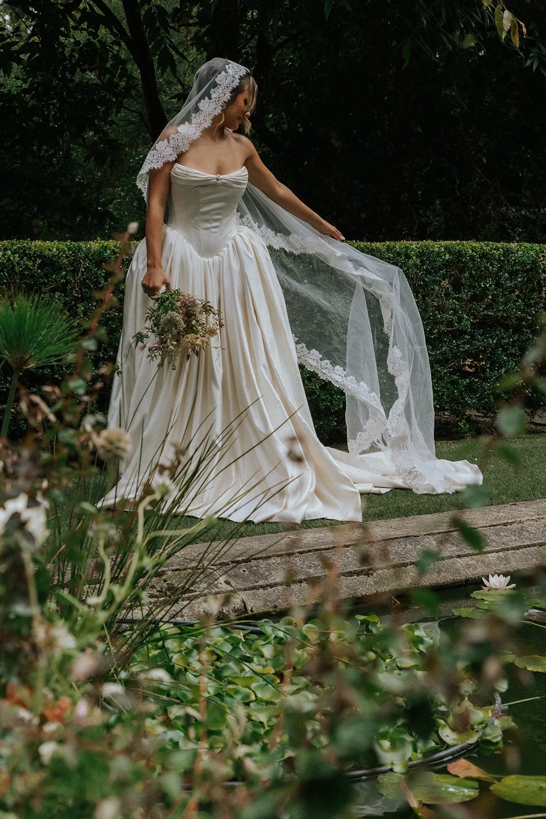 A bride in a white strapless wedding gown and lace veil standing outdoors near a pond with lilies, holding a bouquet of flowers and adjusting her veil.