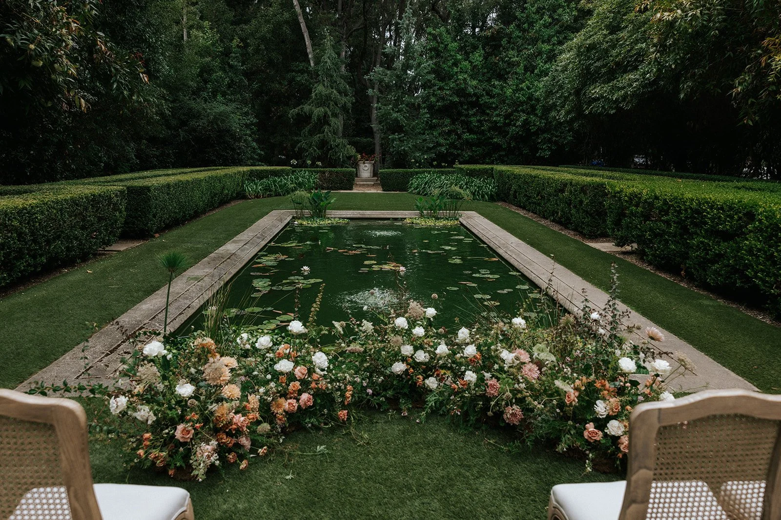 A rectangular garden pond with water lilies, surrounded by flower beds, manicured lawns, and trimmed hedges in a lush green garden. There are two chairs in the foreground facing the pond.