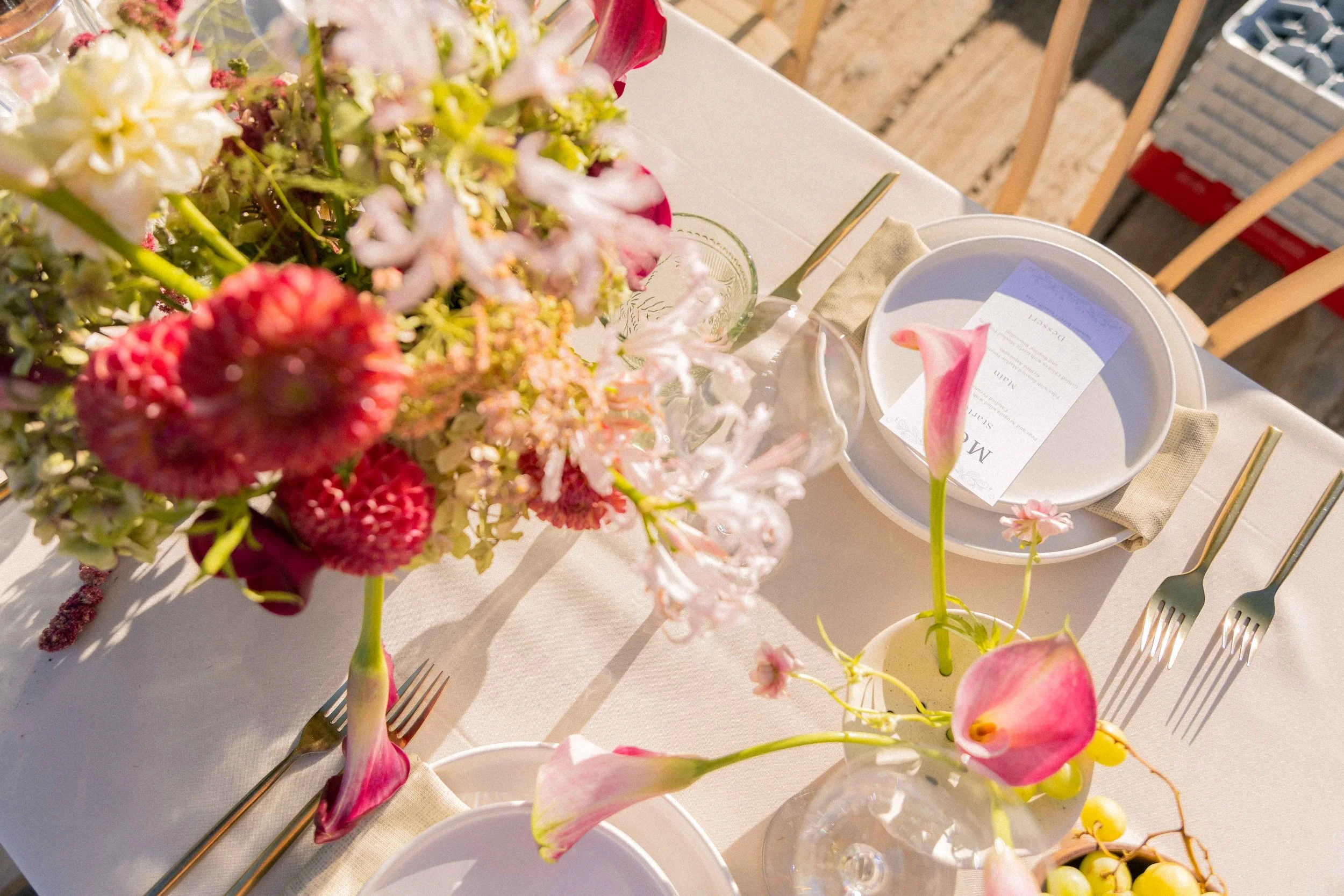 A close-up of a dining table setting with floral arrangements, plates, and utensils, illuminated by sunlight.