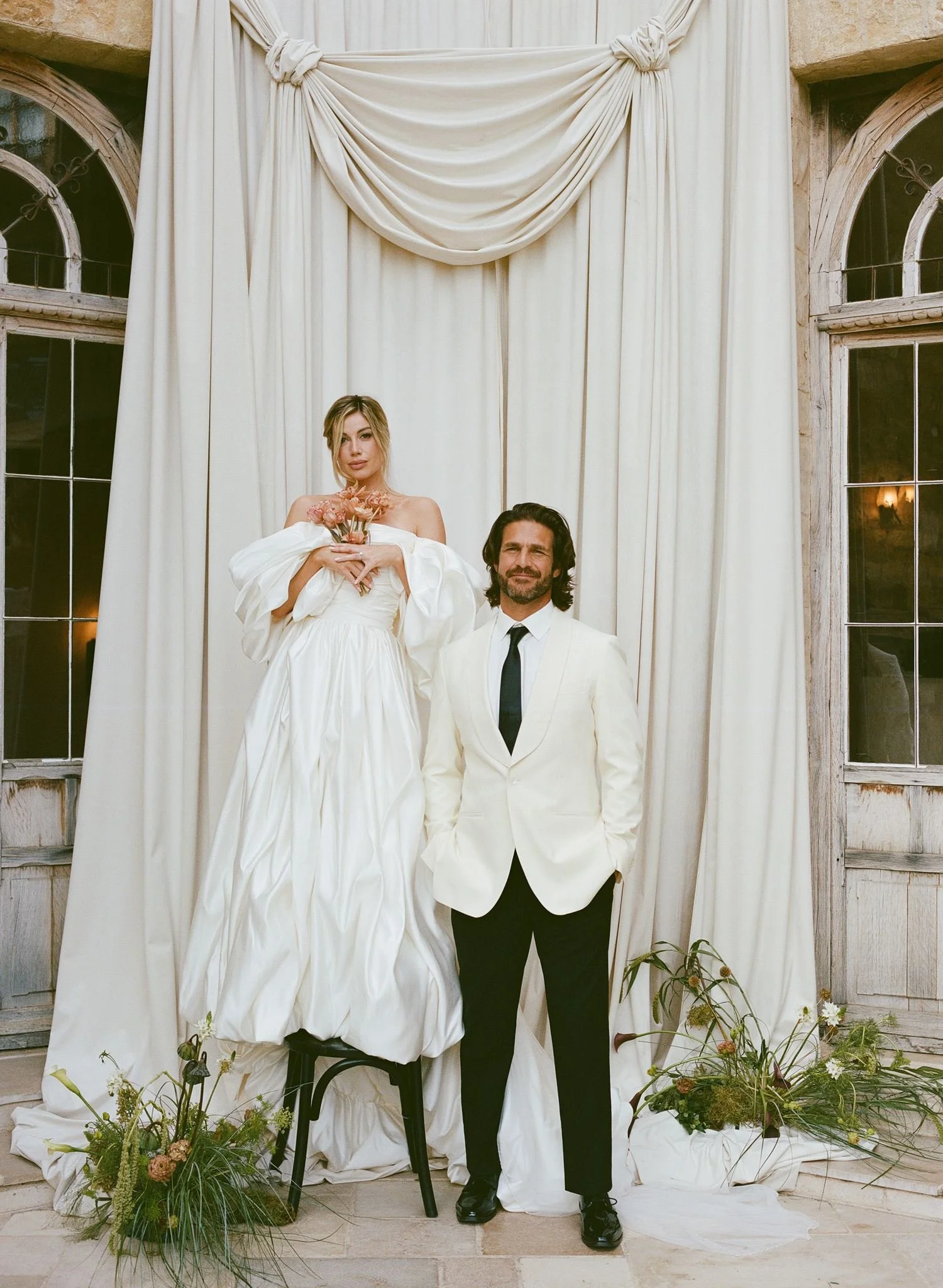 A woman in a white dress holding pink flowers sitting on a chair next to a man in a white suit and black tie standing in front of a white curtain backdrop with windows on either side, surrounded by floral arrangements.