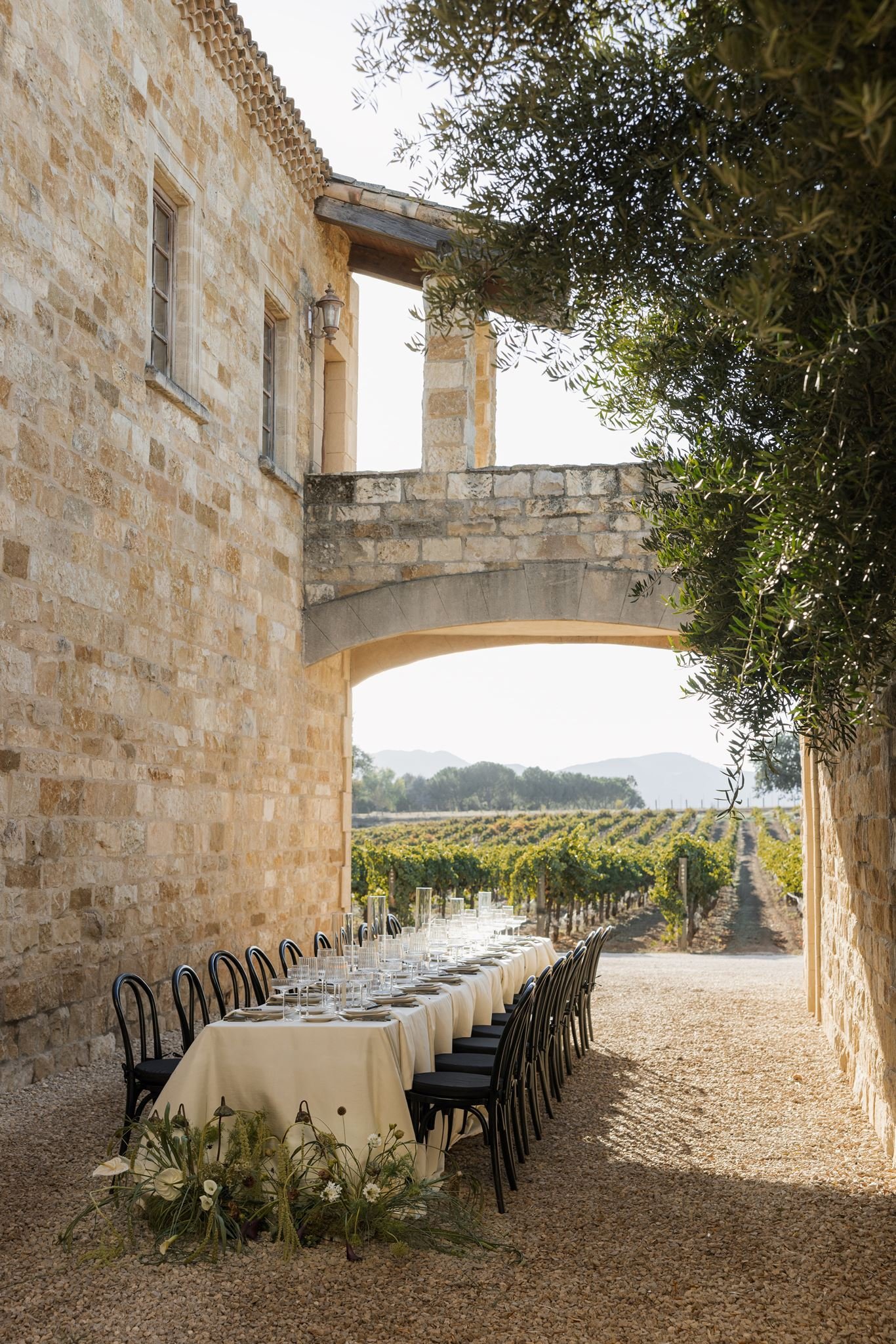 Long dining table set with white tablecloth and glassware sits outdoors under arched stone structure, overlooking vineyards in the background.