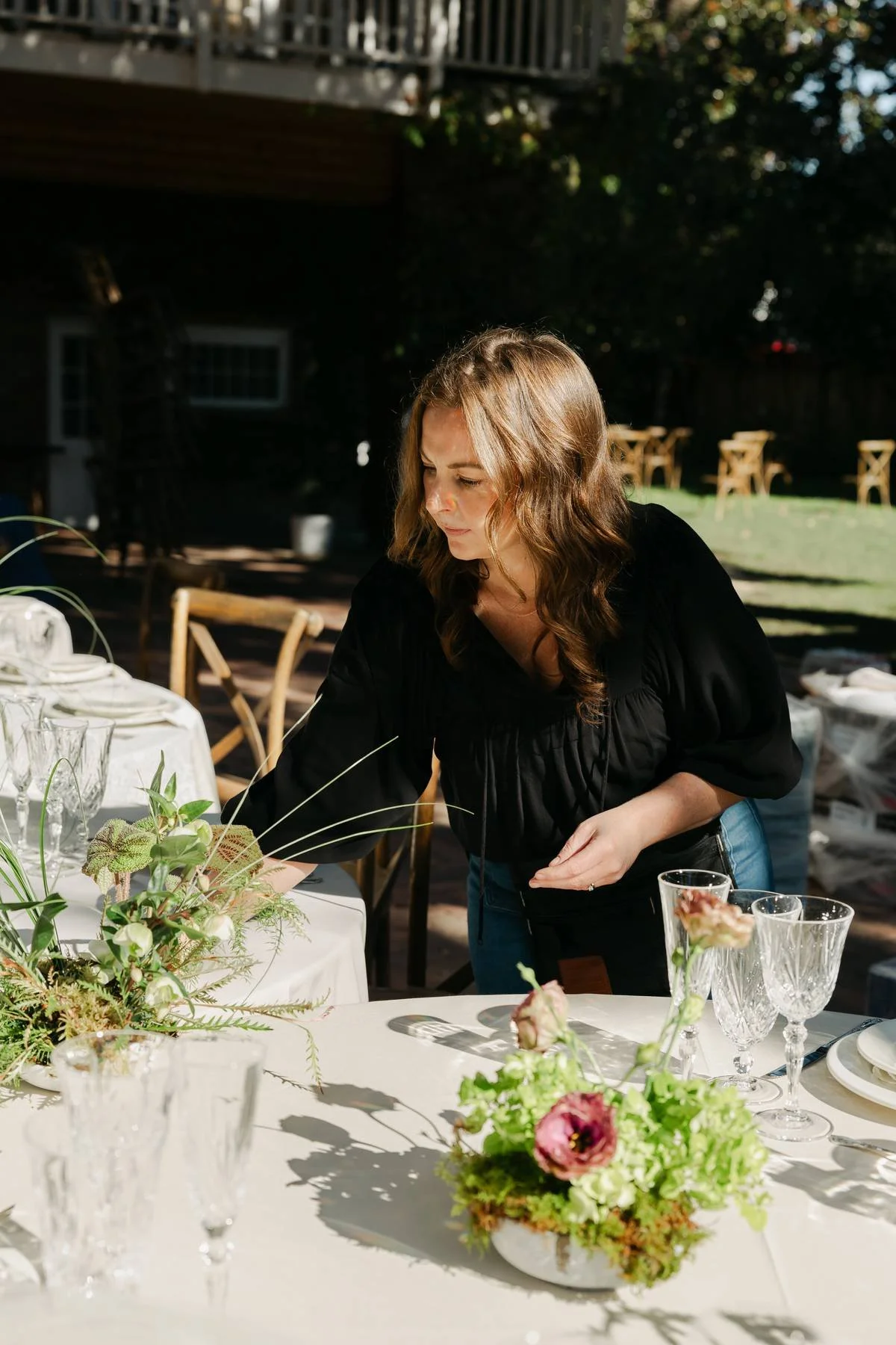 Woman with wavy brown hair arranging floral centerpiece on a decorated table outdoors