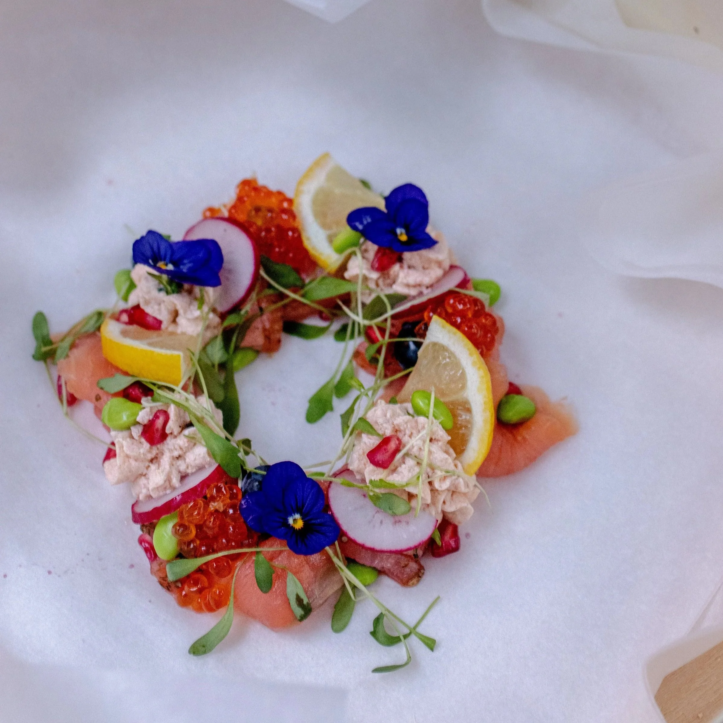 An artistic display of edible flowers, lemon slices, radish slices, salmon, and red and orange fish eggs on a white surface.