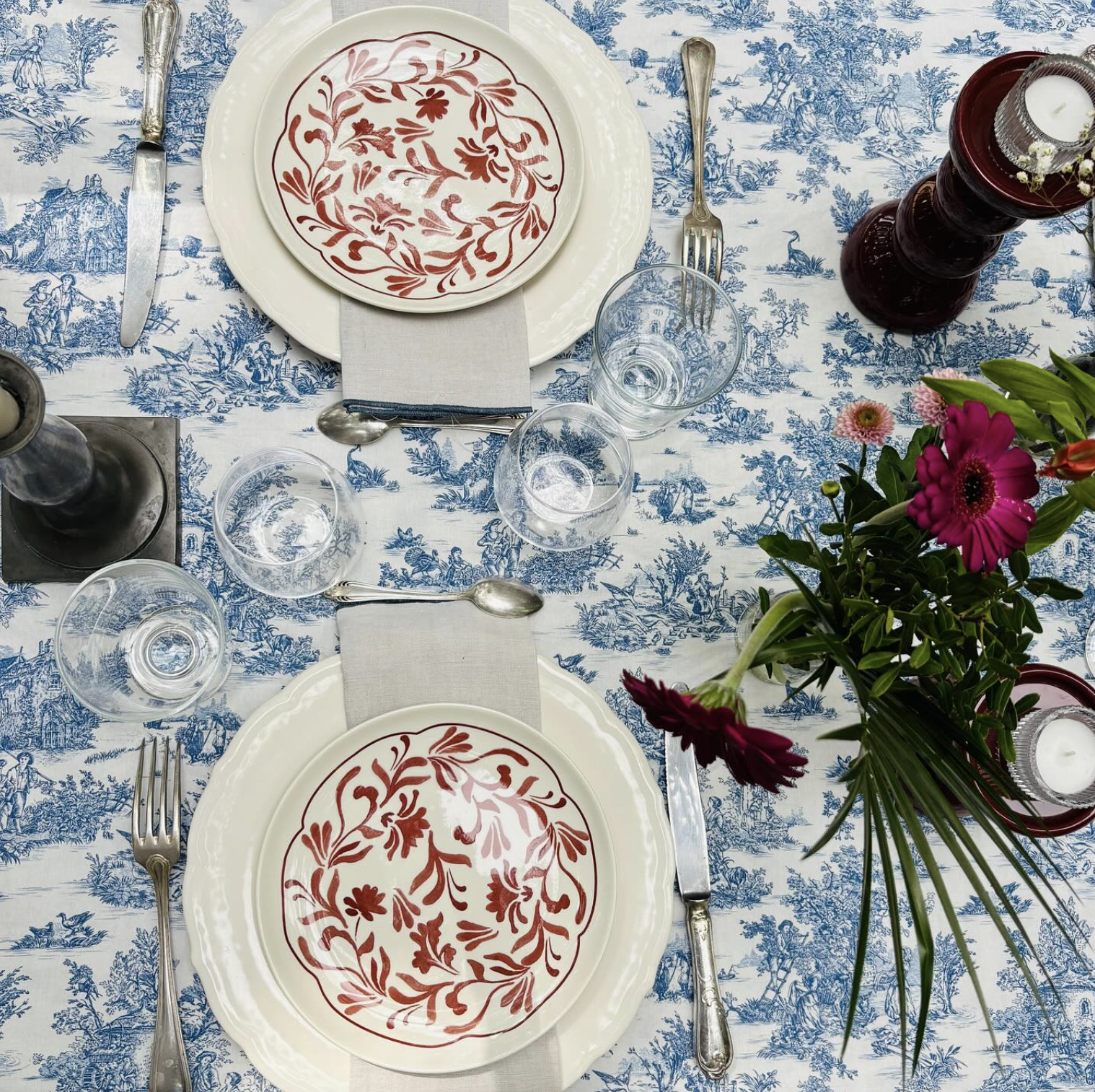 Dinner for two on blue toile linen, with red floral plates, candlesticks and a bunch of red flowers.