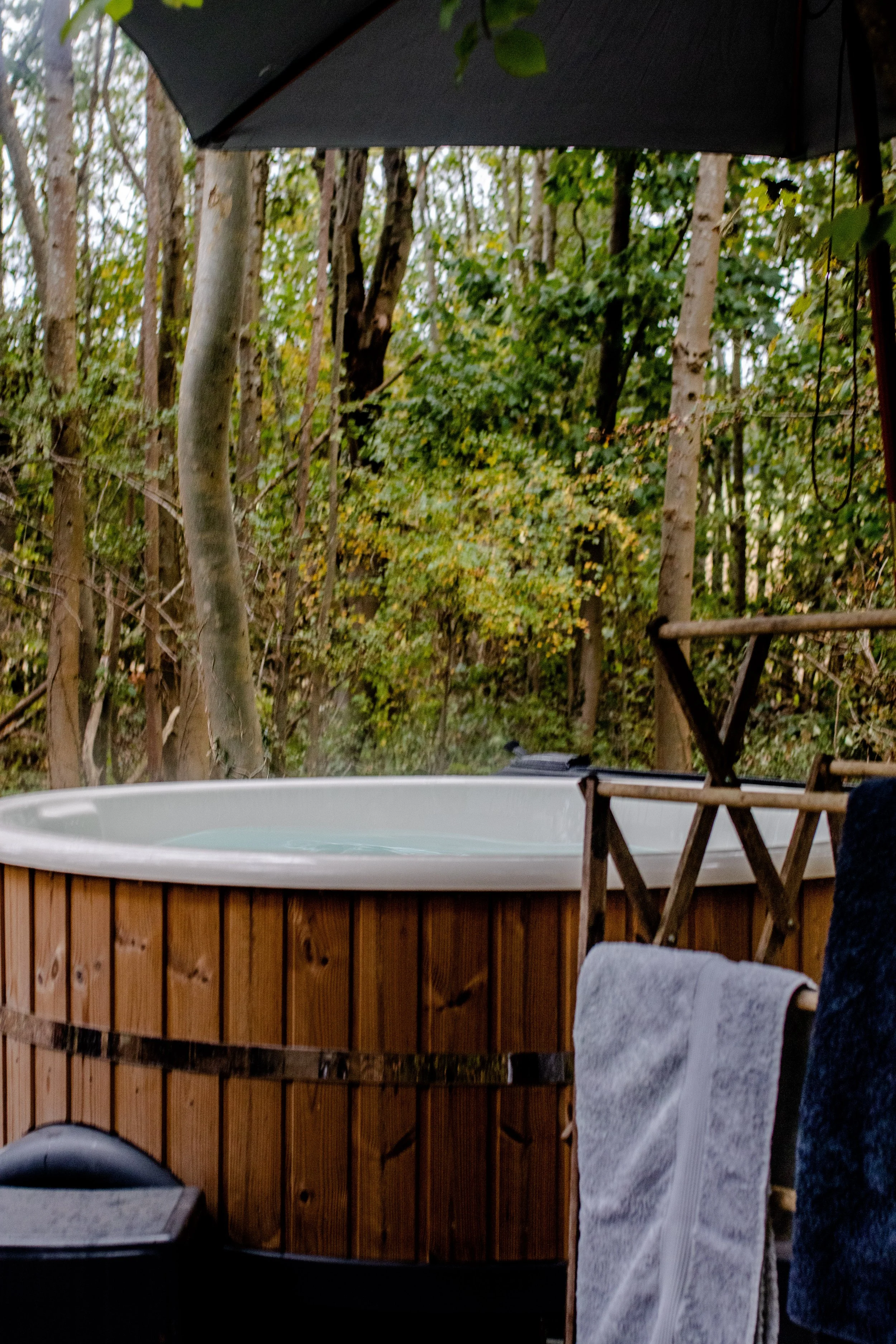 Wooden hot tub with a white interior, positioned outdoors in a wooded area with trees and greenery, partially covered by a black umbrella.