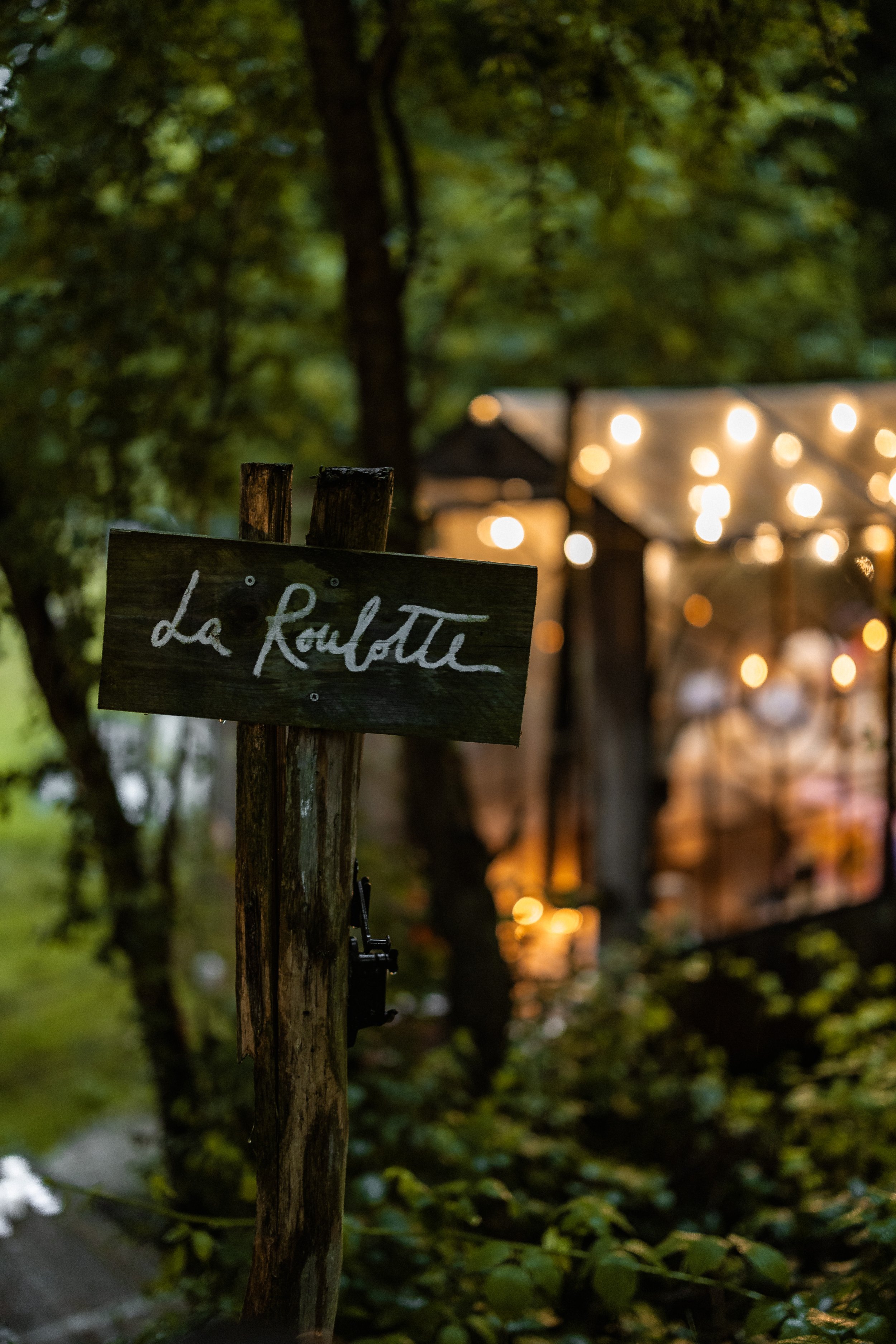 La Roulotte sign at night, with the glass cabin glowing with string lights behind it.