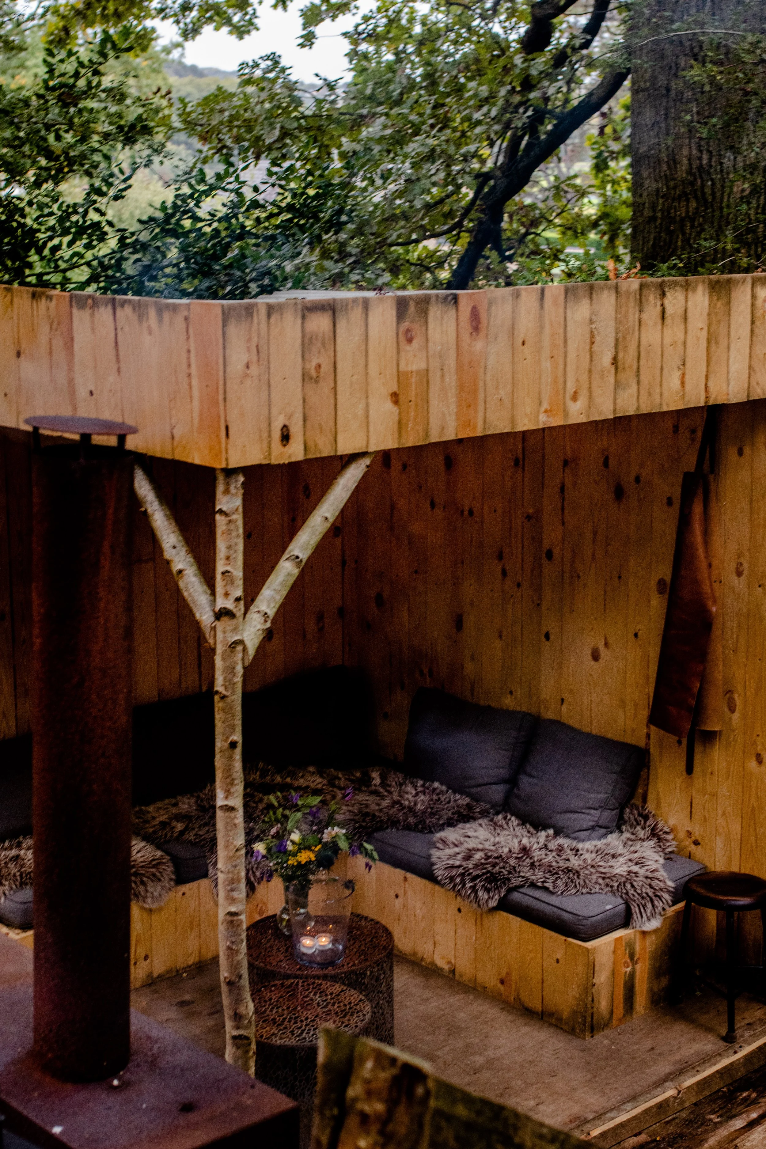 Outdoor seating area with a wooden wall, a black cushioned bench with throw pillows and a fur blanket, a small round table with flower arrangement, and candles, surrounded by trees and greenery.