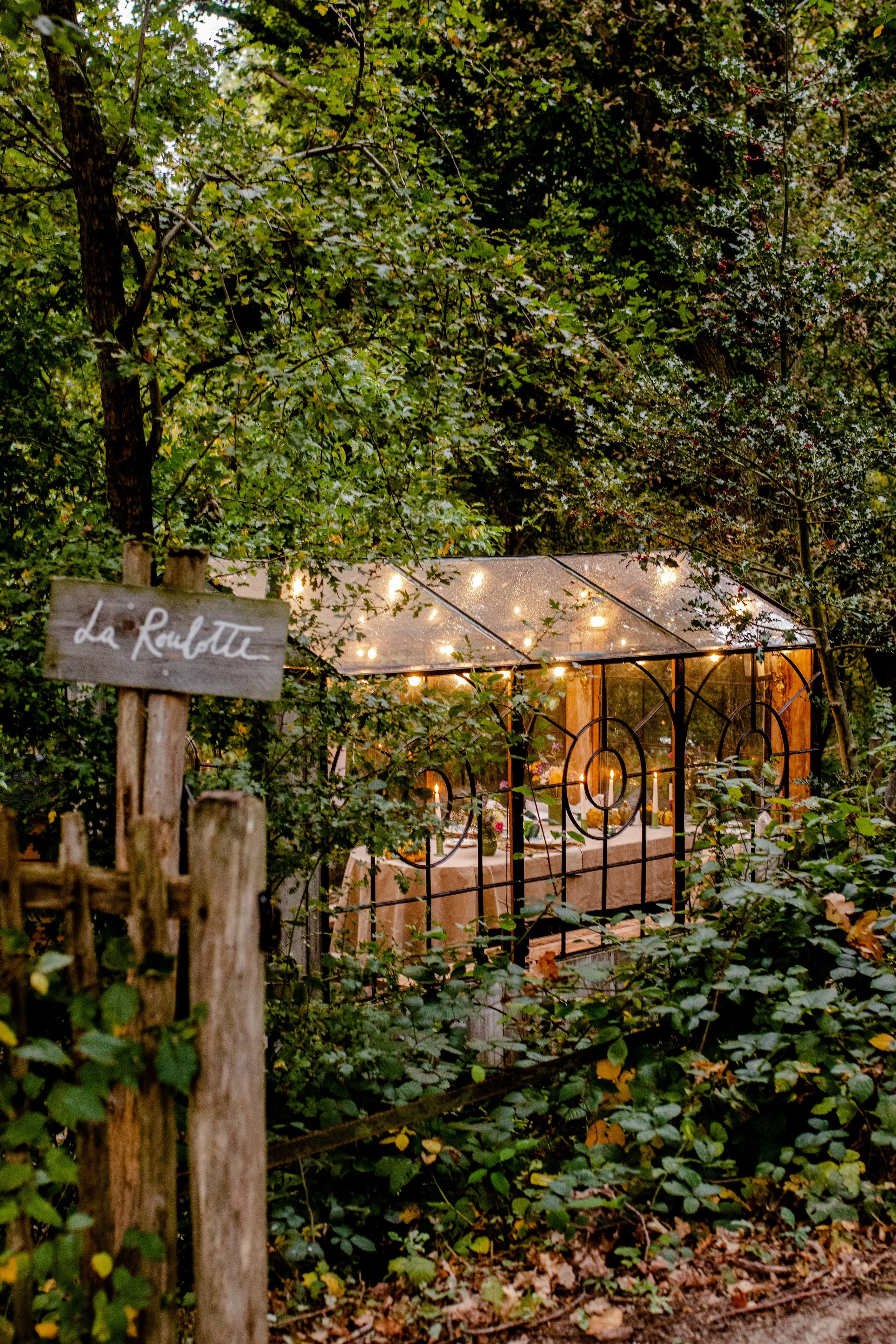La Roulotte glass cabin lit with string lights in the forest near Wavre, with its wooden sign at the entrance.