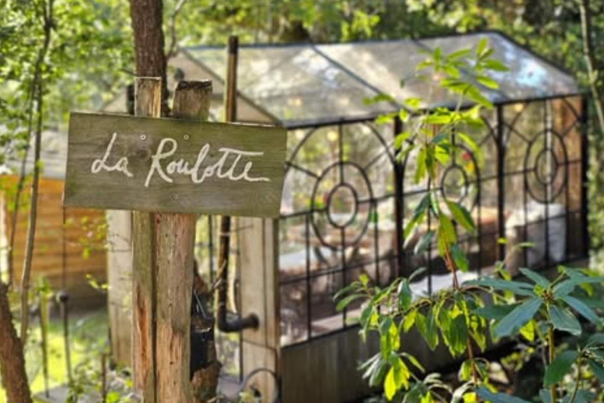 Daytime view of the glass-and-iron cabin behind its handwritten La Roulotte sign, in the woods near Wavre.