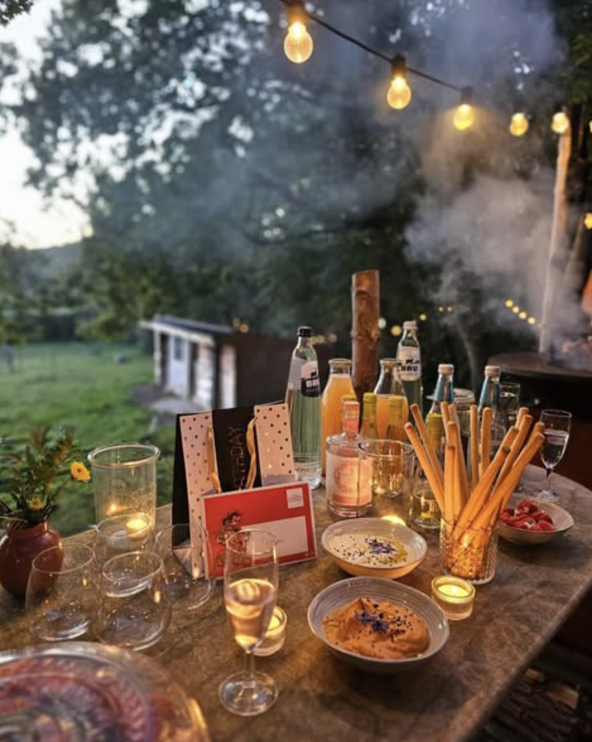 Outdoor table with apéro snacks and drinks under string lights, as the grill heats up in the forest.