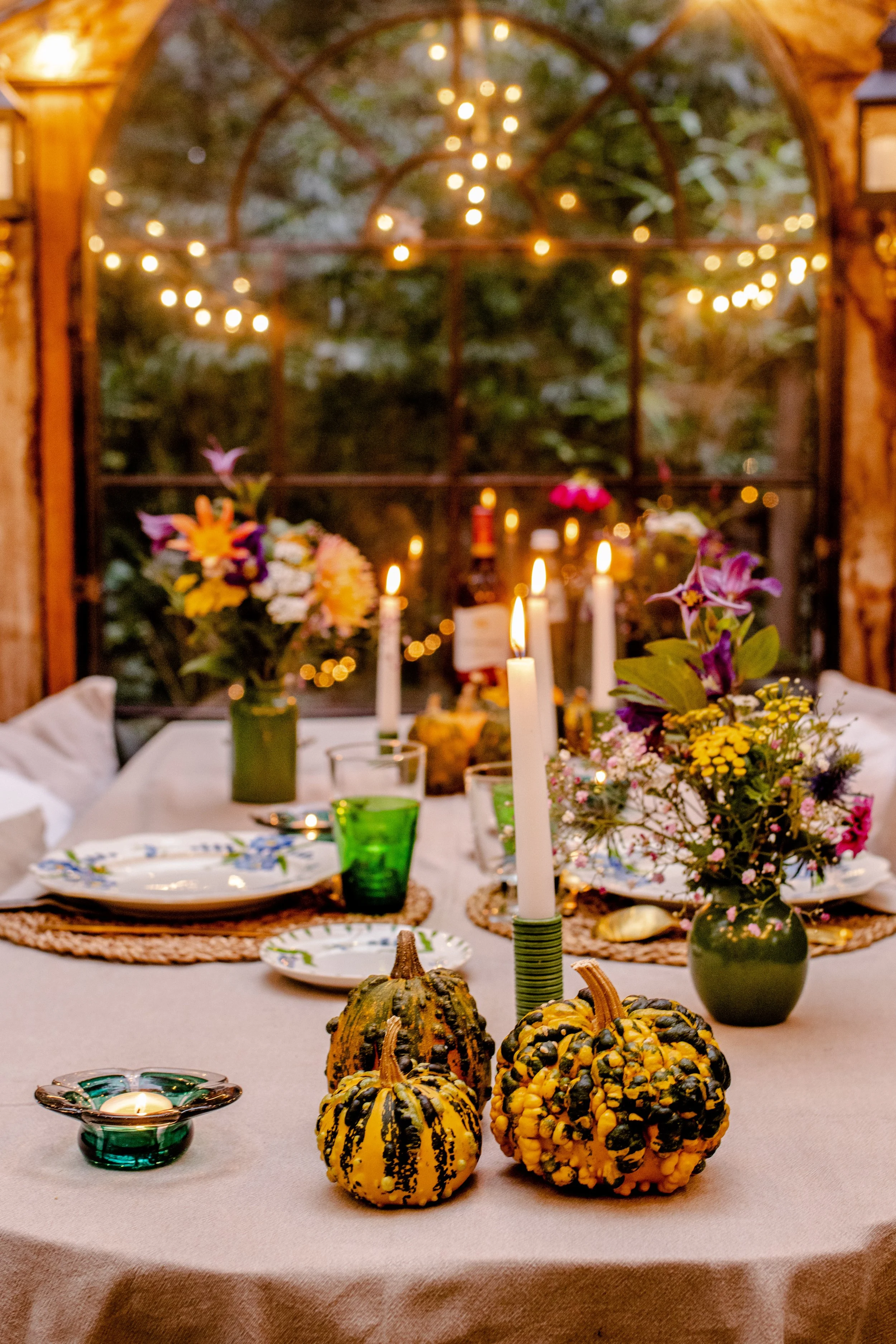 A decorated dining table with autumn-themed gourds, floral arrangements, and candles, set near a window with string lights outside.