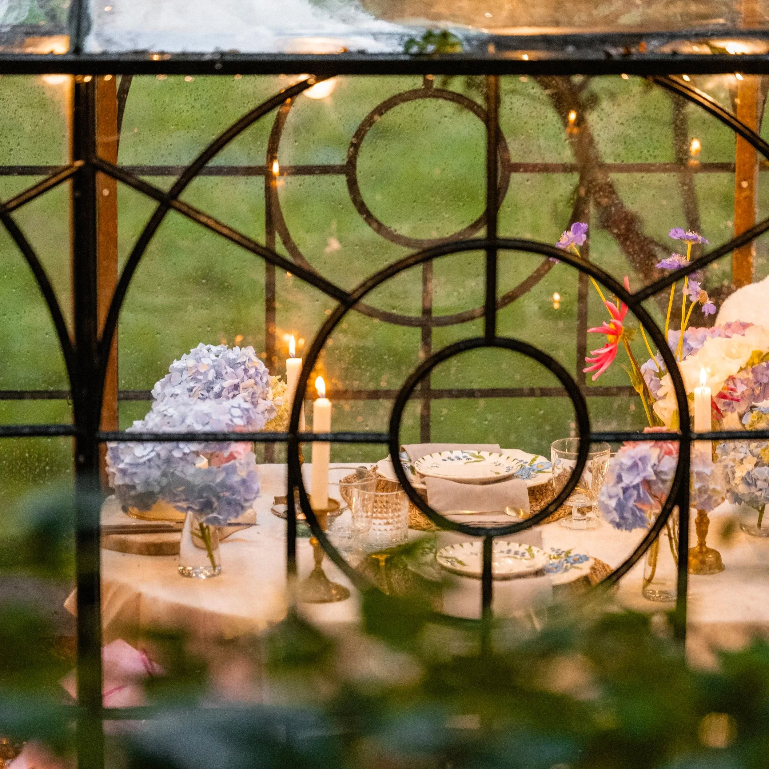 A view through a rain-covered window pane showing a dining table with various plates, glasses, candles, and floral arrangements in an outdoor setting during the evening.