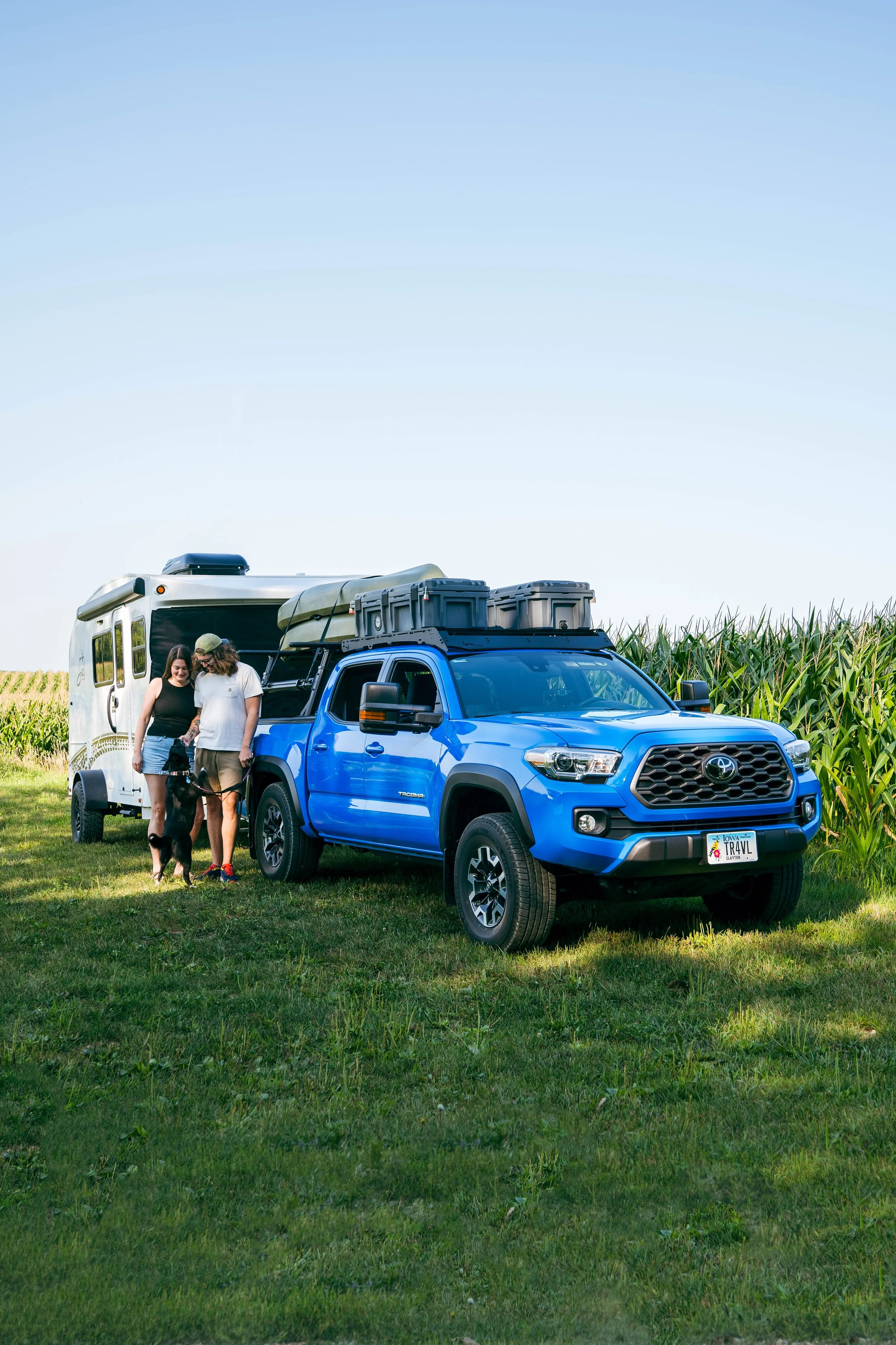 A blue Toyota Tacoma truck parked on a grassy field near a camper trailer, with two people and a dog standing beside the truck.