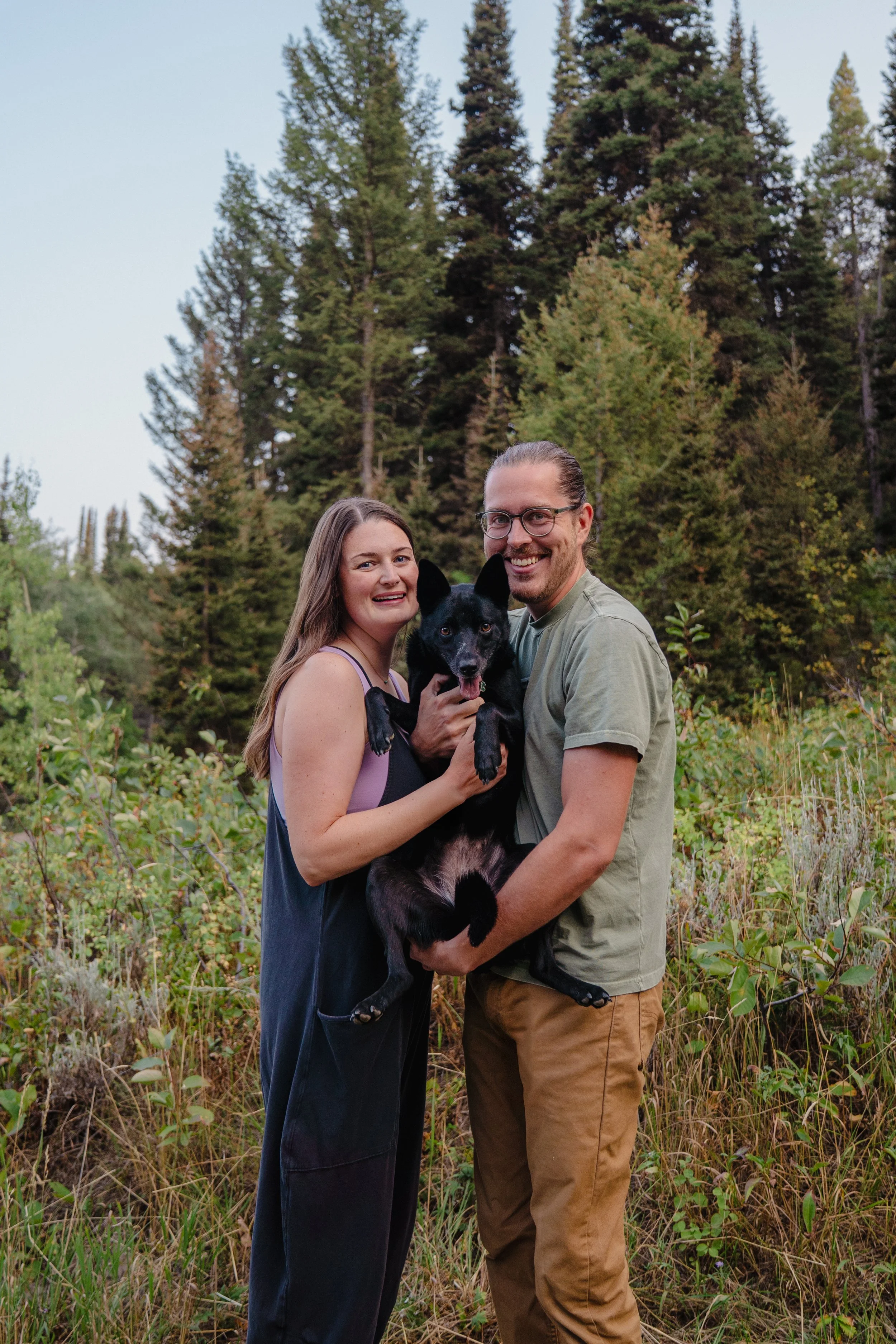 A happy couple with a black dog standing outdoors in a lush green forested area.