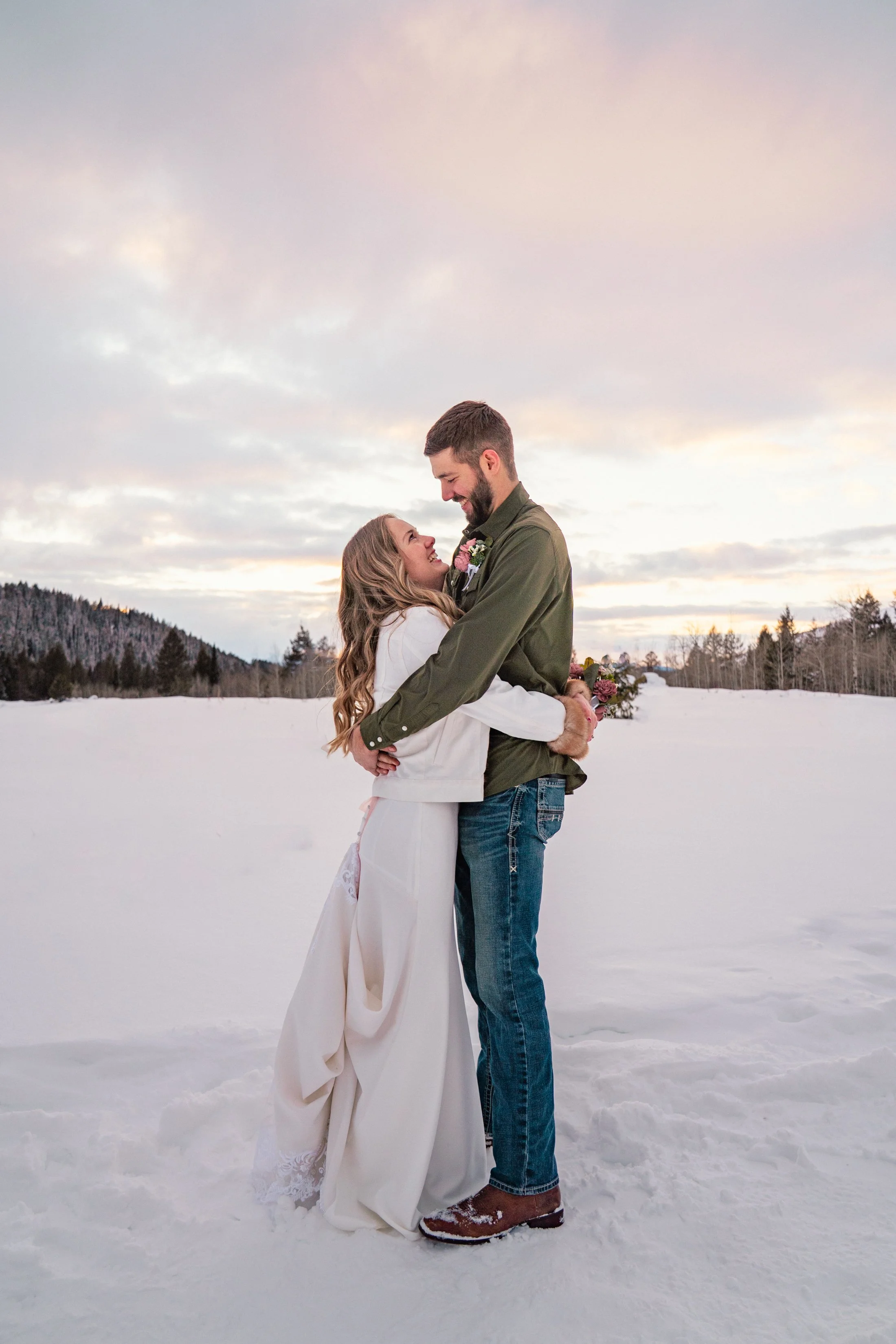 A couple dressed in wedding attire embracing outdoors in a snowy landscape with trees and mountains in the background during sunset.
