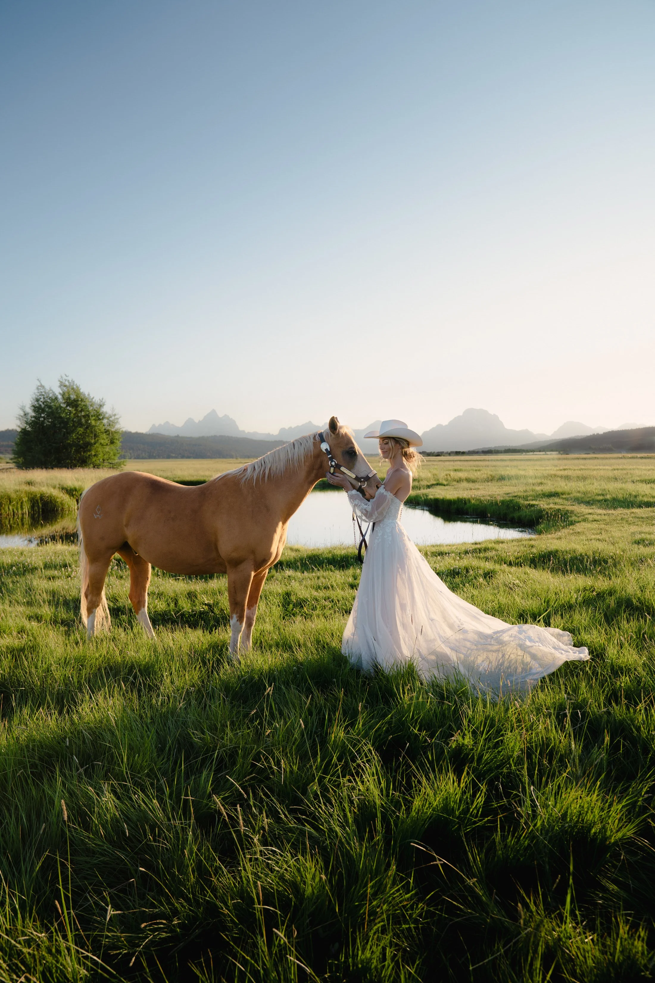 A woman in a white wedding dress and cowboy hat stands next to a light brown horse in a lush green field near a small pond with mountains in the distance during the daytime.
