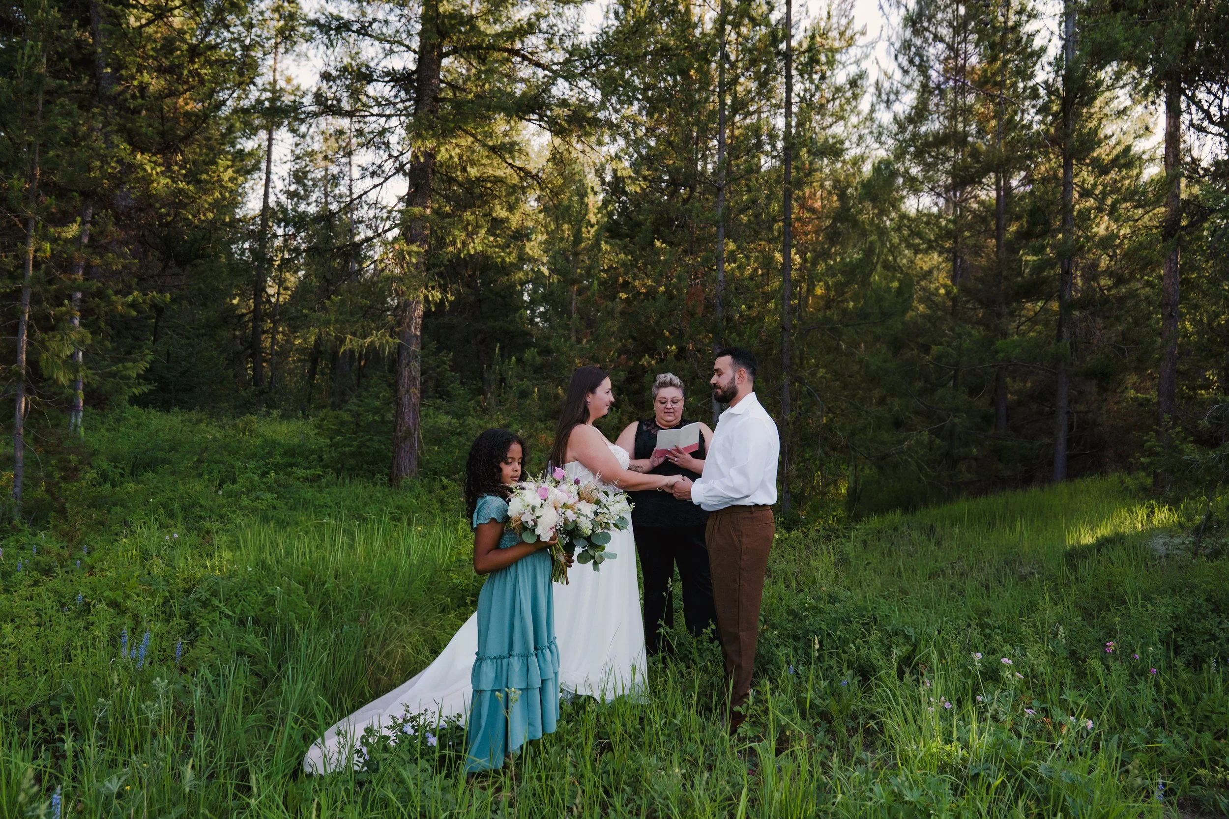A couple getting married in a forested outdoor setting with two young girls, one holding a bouquet and the other standing beside them.