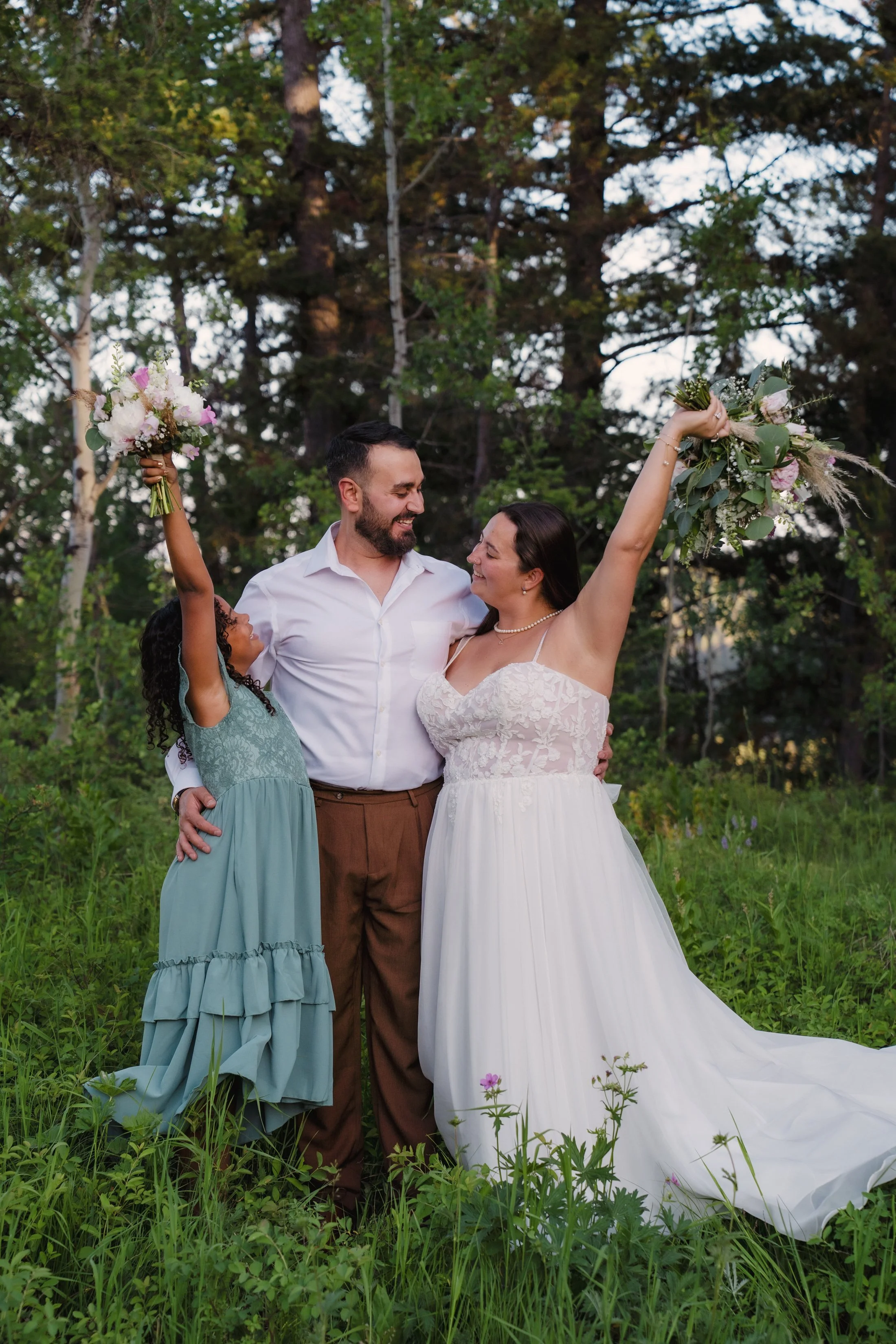 A bride in a white wedding dress and gown holds a large bouquet of flowers.Wedding couple standing outdoors among green grass and trees, smiling at each other, the bride with one arm raised holding a bouquet.