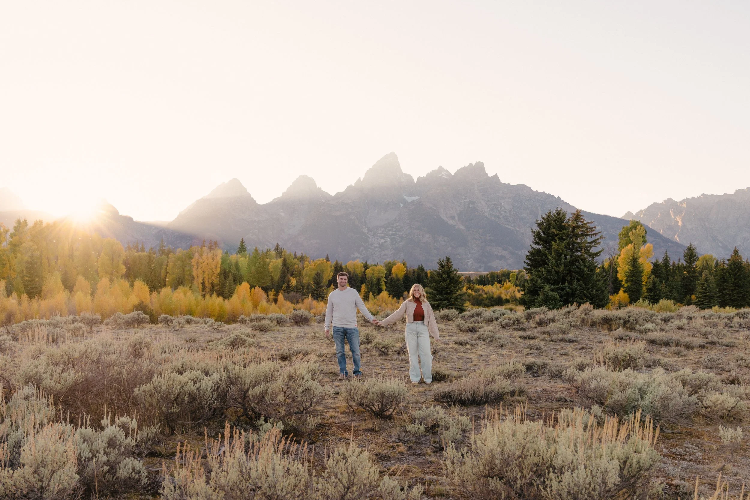 A couple walking hand in hand through a field at sunset with mountains and trees in the background at Schwabacher Landing.