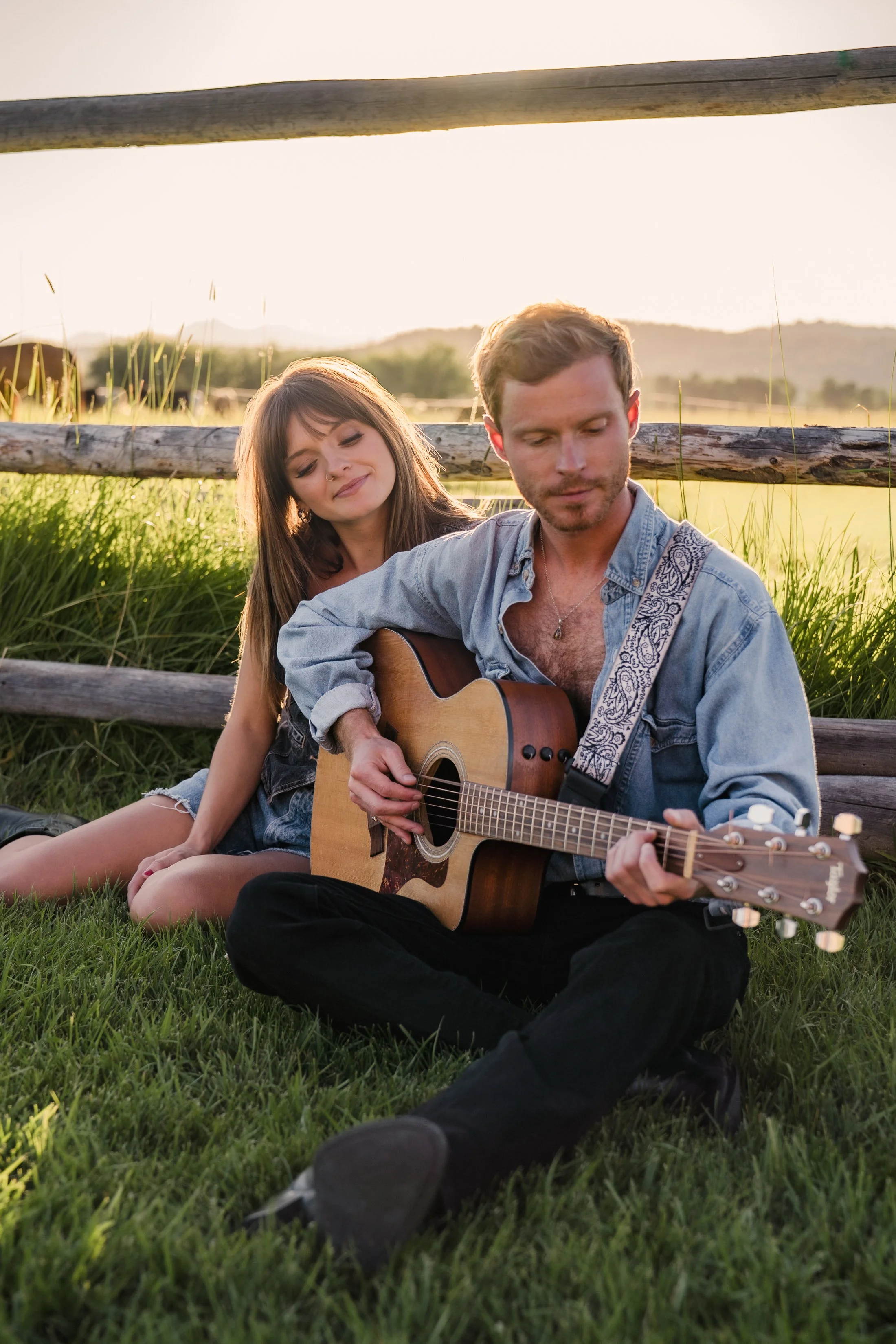 A young man playing an acoustic guitar while sitting on the grass with a young woman leaning on his shoulder, outdoors in a field with a wooden fence and mountains in the background during sunset.