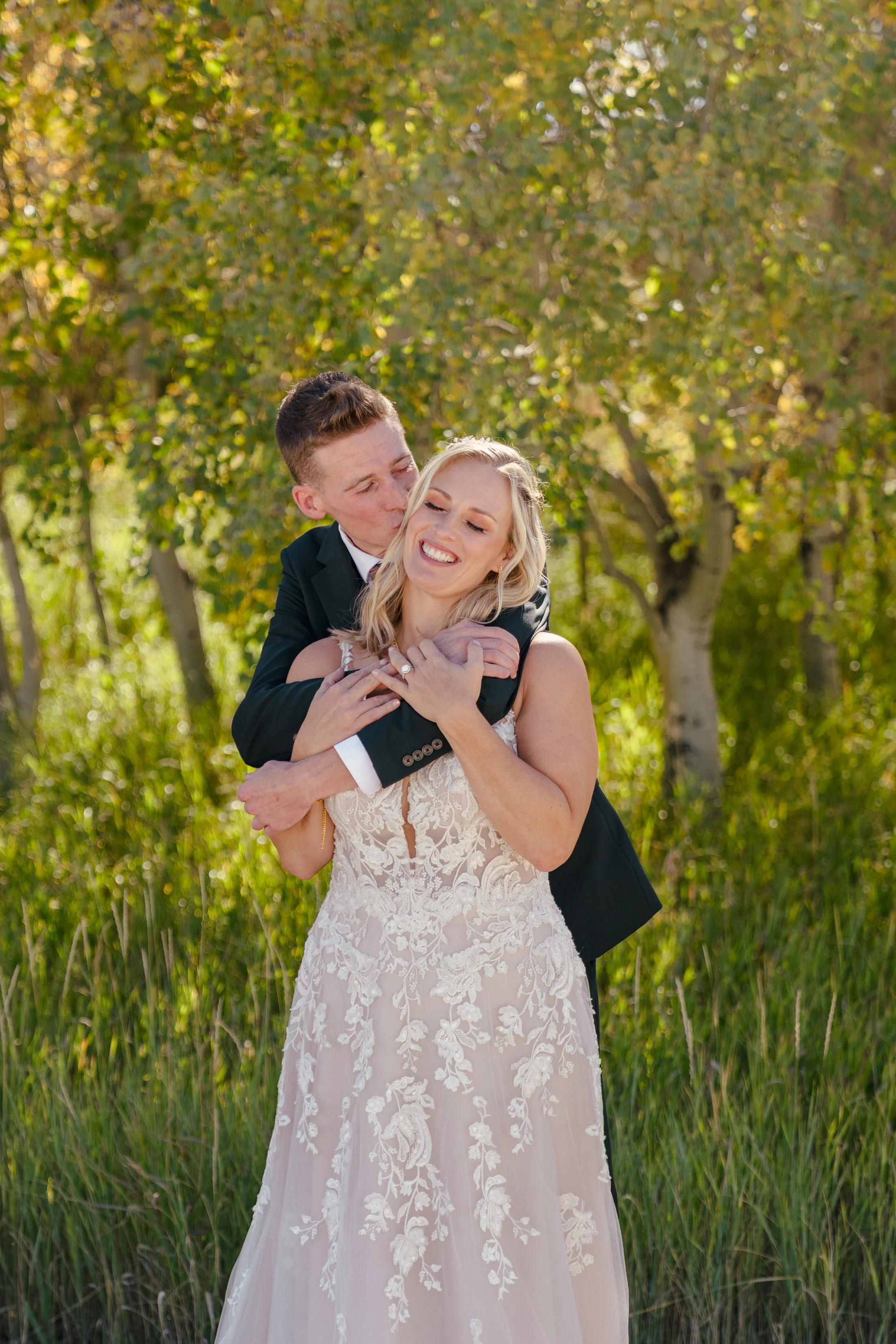 A smiling woman in a lace wedding dress is being hugged from behind by a man in a black suit. They are outdoors in a field with green grass and trees with yellow and green leaves, suggesting autumn in Teton Valley.