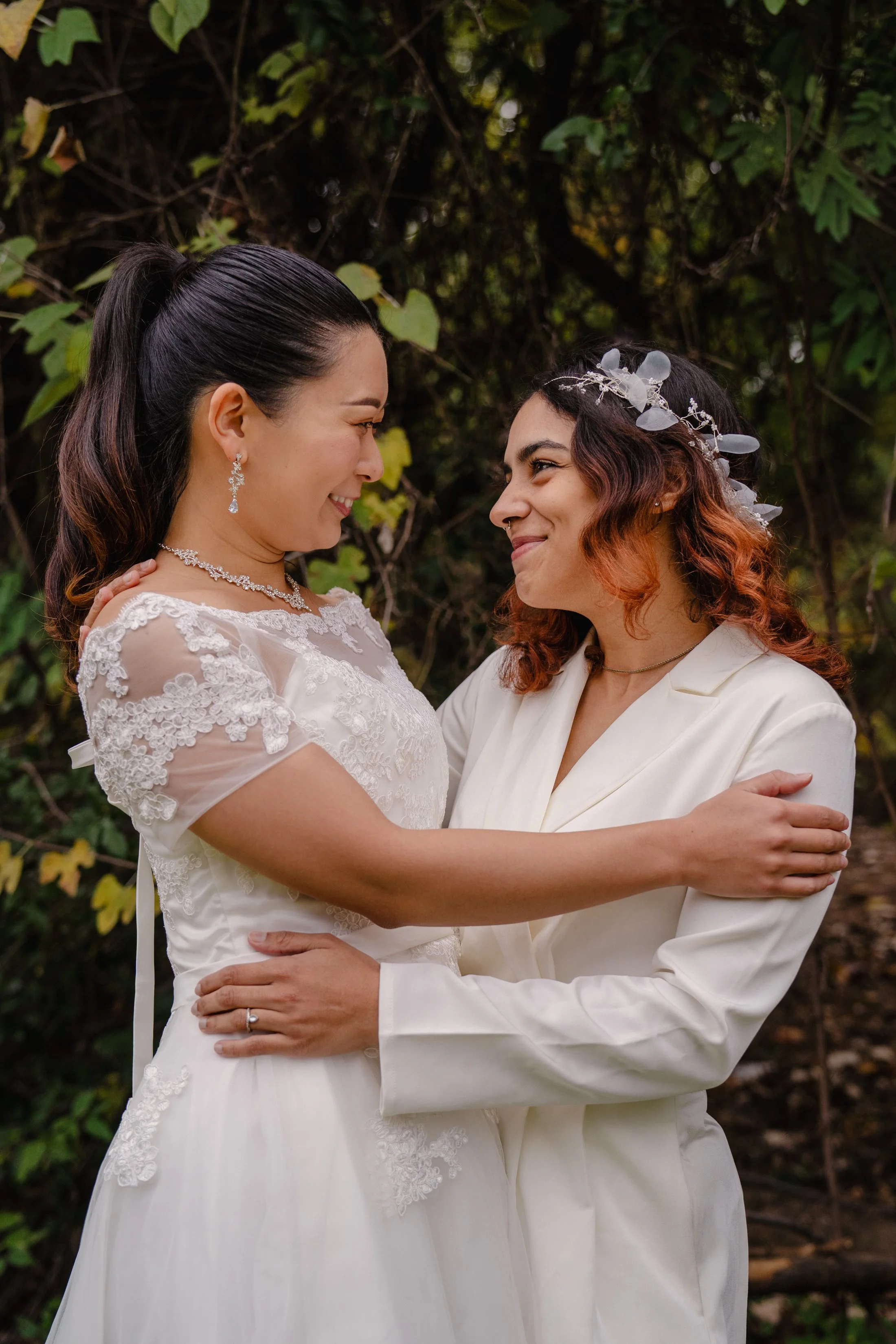 Two women in wedding attire hugging and smiling at each other outdoors surrounded by greenery.