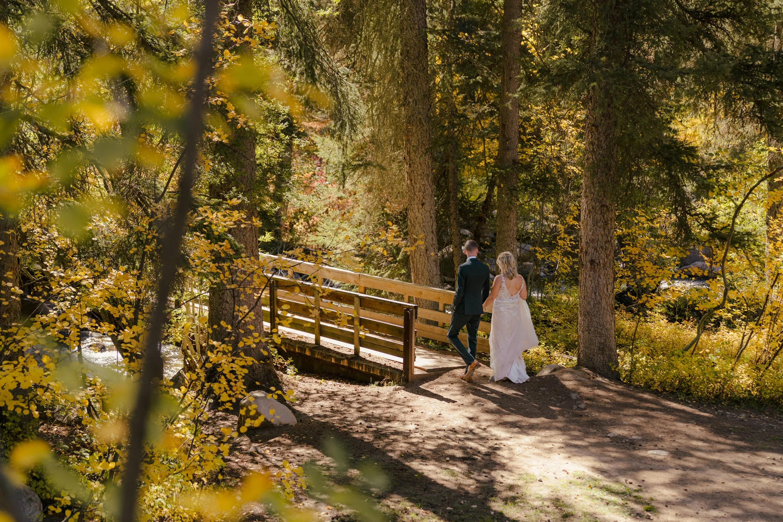 A bride and groom walking on a wooden bridge in a forest with tall trees and yellow leaves during fall.